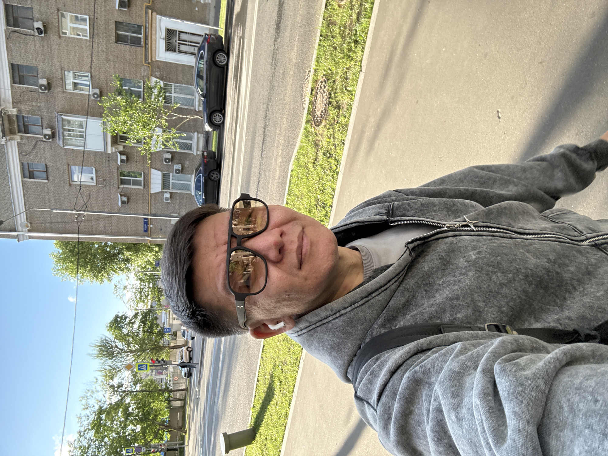 A selfie taken from a low angle shows a person wearing sunglasses and a grey hoodie with a zipper. The person has short dark hair and is wearing white wireless earbuds. The background depicts a street scene in Moscow, Russia, with a multi-story apartment building, trees, cars, and a street. The building is made of brick with multiple windows and air conditioning units. Two cars, a dark sedan and a dark SUV, are parked on the street. The sky is blue with some clouds, and the sun is casting shadows. A traffic light with a pedestrian crossing signal is visible in the distance. A trash can is also present on the sidewalk.