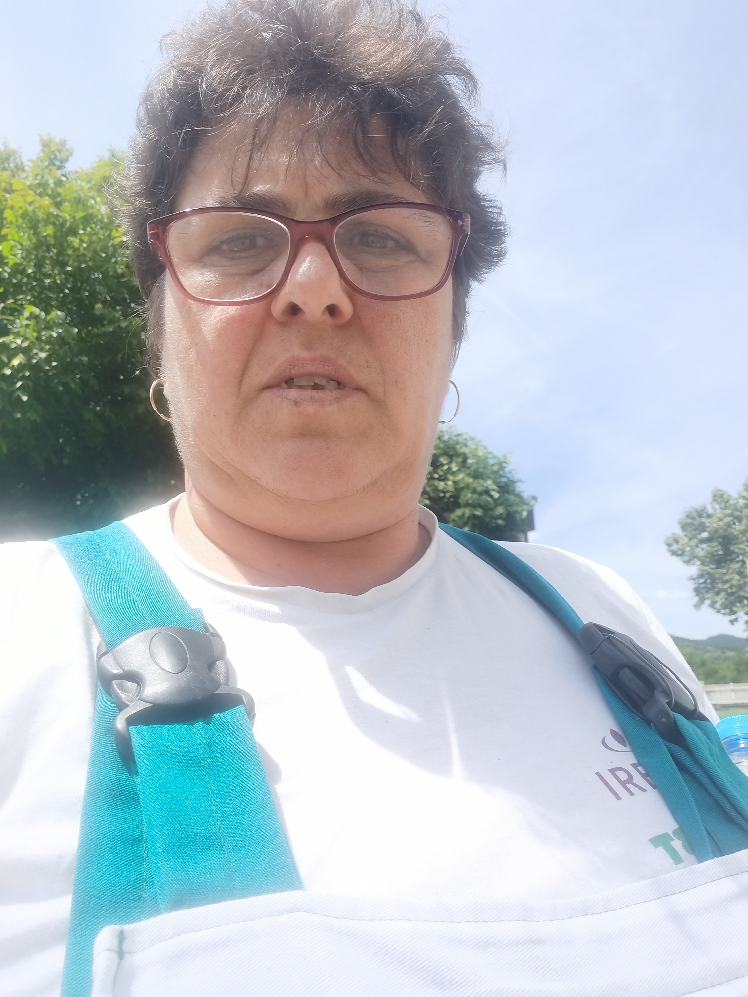 This is a close-up selfie of a middle-aged woman with curly, dark hair and glasses. She is wearing a white t-shirt with a teal strap over her shoulder, possibly part of a bag or harness. A black buckle is visible on the strap. The woman has a neutral expression.
The background is a bright, sunny sky with some green trees on the left and right sides, suggesting an outdoor setting. The overall lighting indicates it is daytime, likely morning or afternoon. The presence of lush greenery hints at a temperate climate and possibly a park or natural environment.
There is some faint text visible on the woman's t-shirt, appearing to be in shades of purple and green, with the letters "IRE" clearly legible.
The image was taken in Lomets, Bulgaria, a fact that is not visually discernible from the photograph itself but is provided as context. The woman's attire and the setting do not specifically indicate any particular event, but she appears to be engaged in an outdoor activity.
