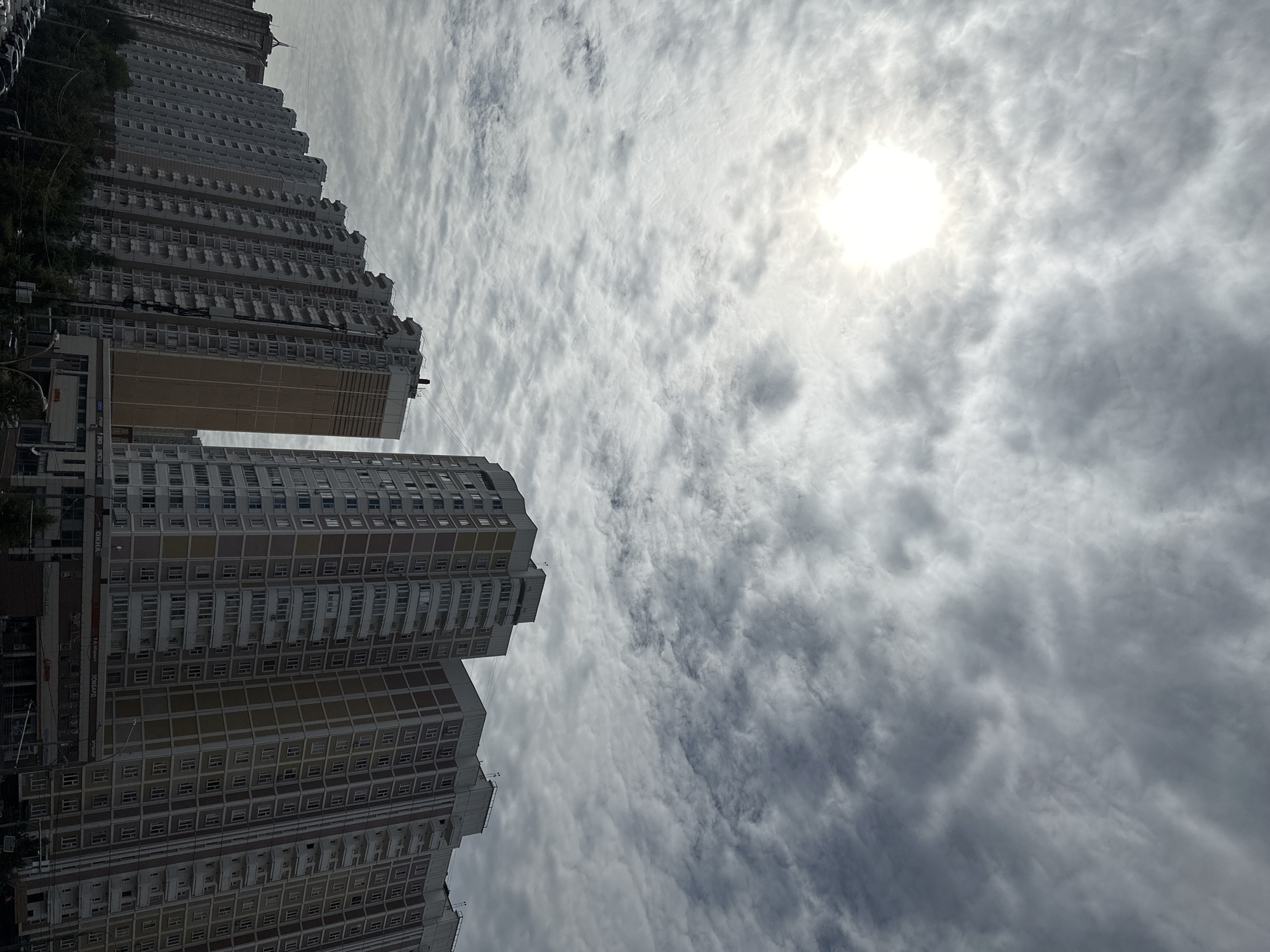 This is a photograph taken from a low angle, looking up at a series of residential apartment buildings in Podolsk, Russia. The sky is overcast with textured clouds, and the sun is visible as a bright, hazy circle in the upper left quadrant. The apartment buildings are tall, multi-story structures with repeating patterns of windows and balconies. They are primarily light-colored with some sections featuring darker accents. The buildings are arranged in a row, receding into the distance. In the foreground, the tops of trees are visible, along with some street-level commercial establishments and parked cars. The scene depicts a typical urban residential area under a cloudy sky.