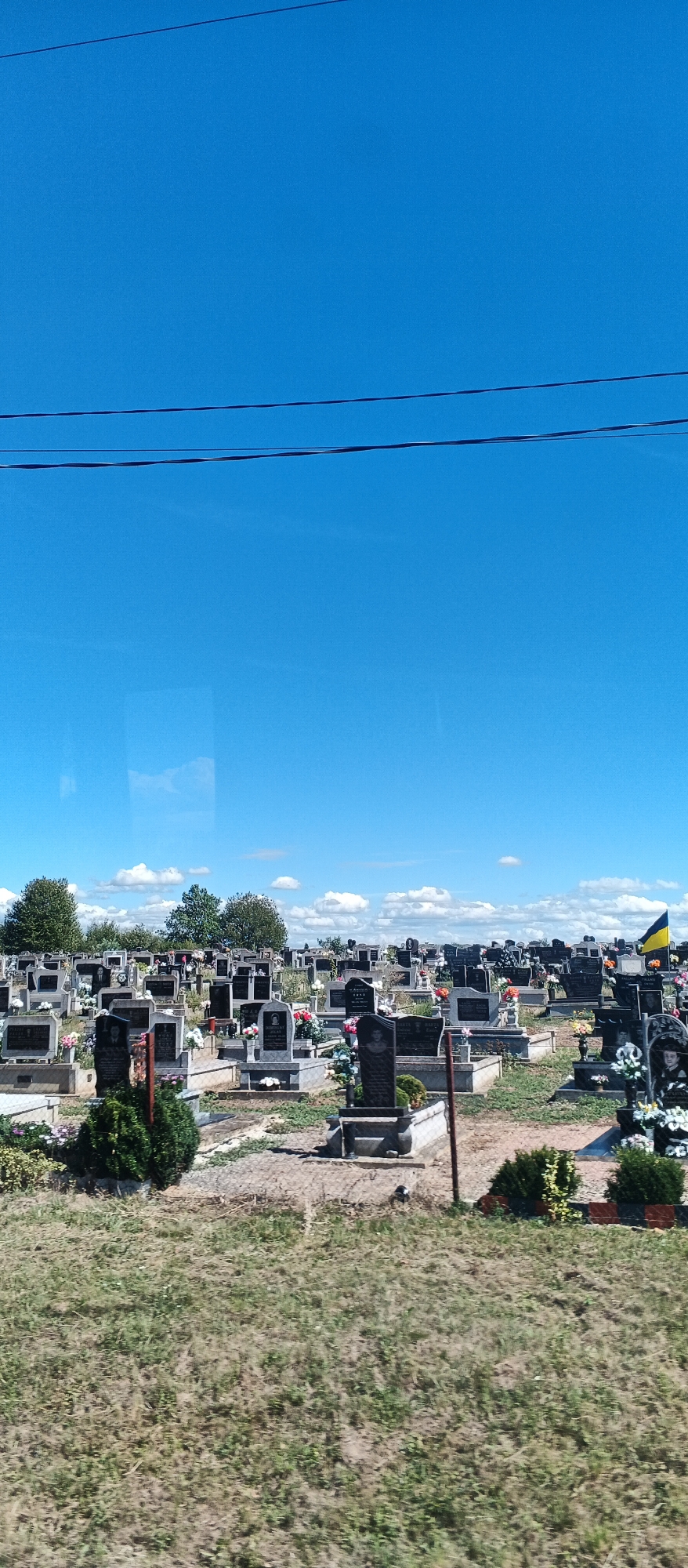 A cemetery is visible under a bright blue sky with scattered white clouds. Numerous gravestones of various sizes and materials are arranged in rows. Some gravestones are adorned with flowers. A Ukrainian flag is visible on a flagpole in the distance. The ground is covered with grass and some sparse vegetation. Power lines are visible in the upper part of the image. The scene appears to be a peaceful and well-maintained cemetery. This cemetery is located in Hat, Ukraine.