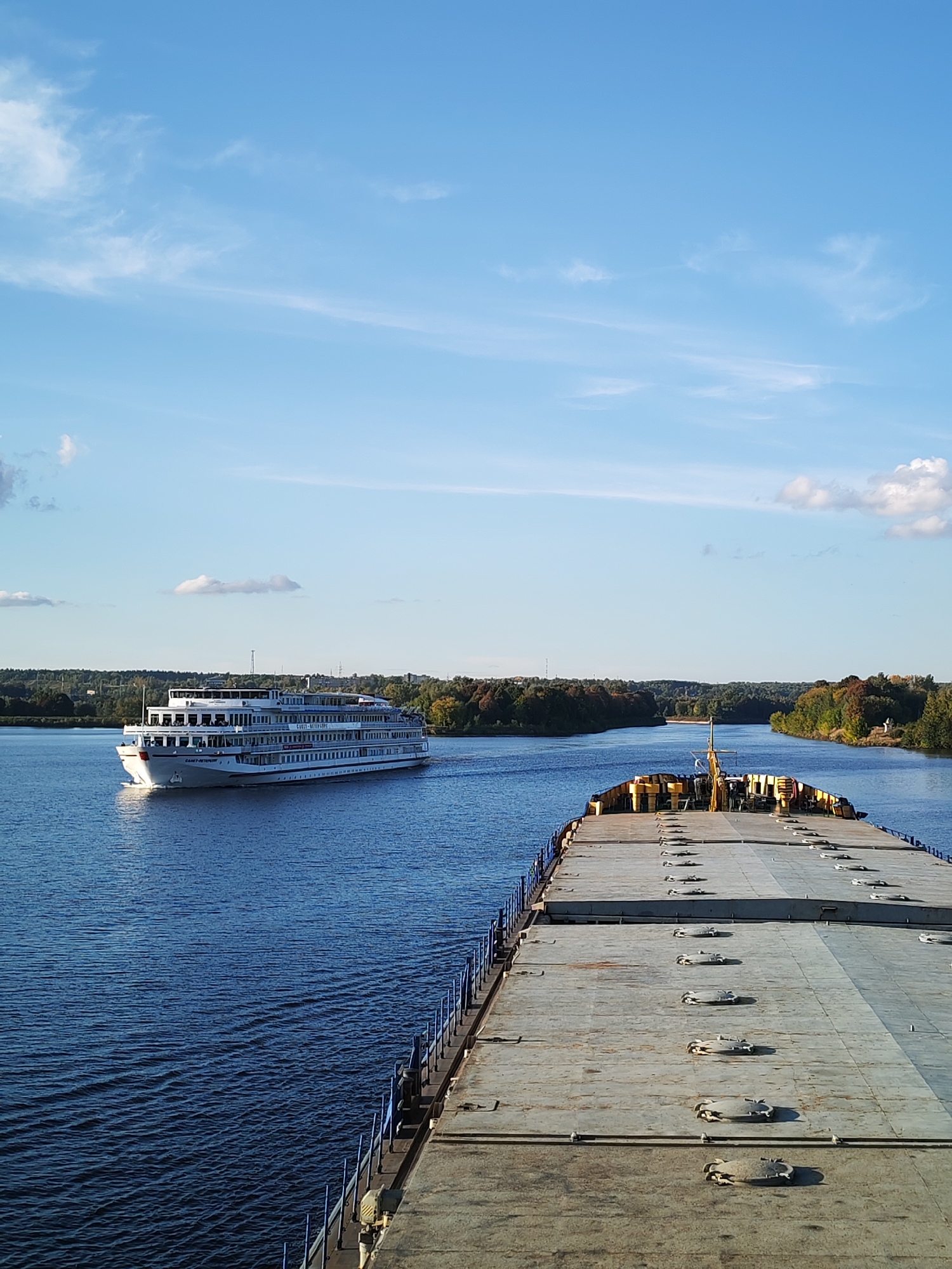This is an outdoor, vertical, eye-level, full shot of a river scene in Letchik-Ispytatel', Russia. The sky is a bright blue with scattered white clouds. The river is a deep blue with gentle ripples on its surface. In the foreground, a flat-bottomed barge is visible, its deck covered with numerous circular hatches and a blue railing along its side. Further in the distance, a large white passenger ship with multiple decks and windows is sailing on the river. The riverbanks are lined with lush green trees, some showing hints of autumn colors. The overall scene depicts a serene day on a wide river with two vessels.