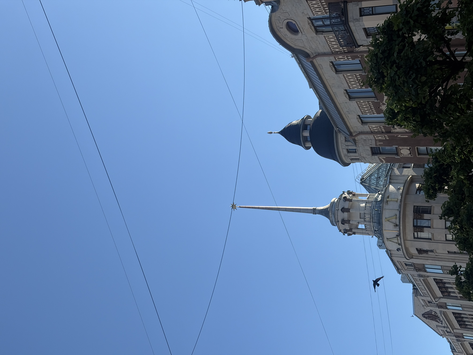 This media file depicts an upward-facing view of architectural landmarks against a clear blue sky. The primary subjects are two distinct buildings with ornate facades, one featuring a dark, domed turret and the other a tall, slender spire topped with a star. Power lines crisscross the sky, adding a modern element to the scene. A lone bird is captured in flight between the buildings.
The scene appears to be set during daytime, indicated by the bright, cloudless sky. The architecture suggests a European urban setting, consistent with the provided location context of Saint Petersburg, Russia. One building has a prominent name displayed on its facade, reading "ИЗВЕСТИЯ" in Cyrillic script. The overall impression is one of grandeur and historical significance, with a sense of open space despite being in an urban environment. There is no discernible activity or event taking place other than the bird's flight. The weather appears to be fair and sunny.