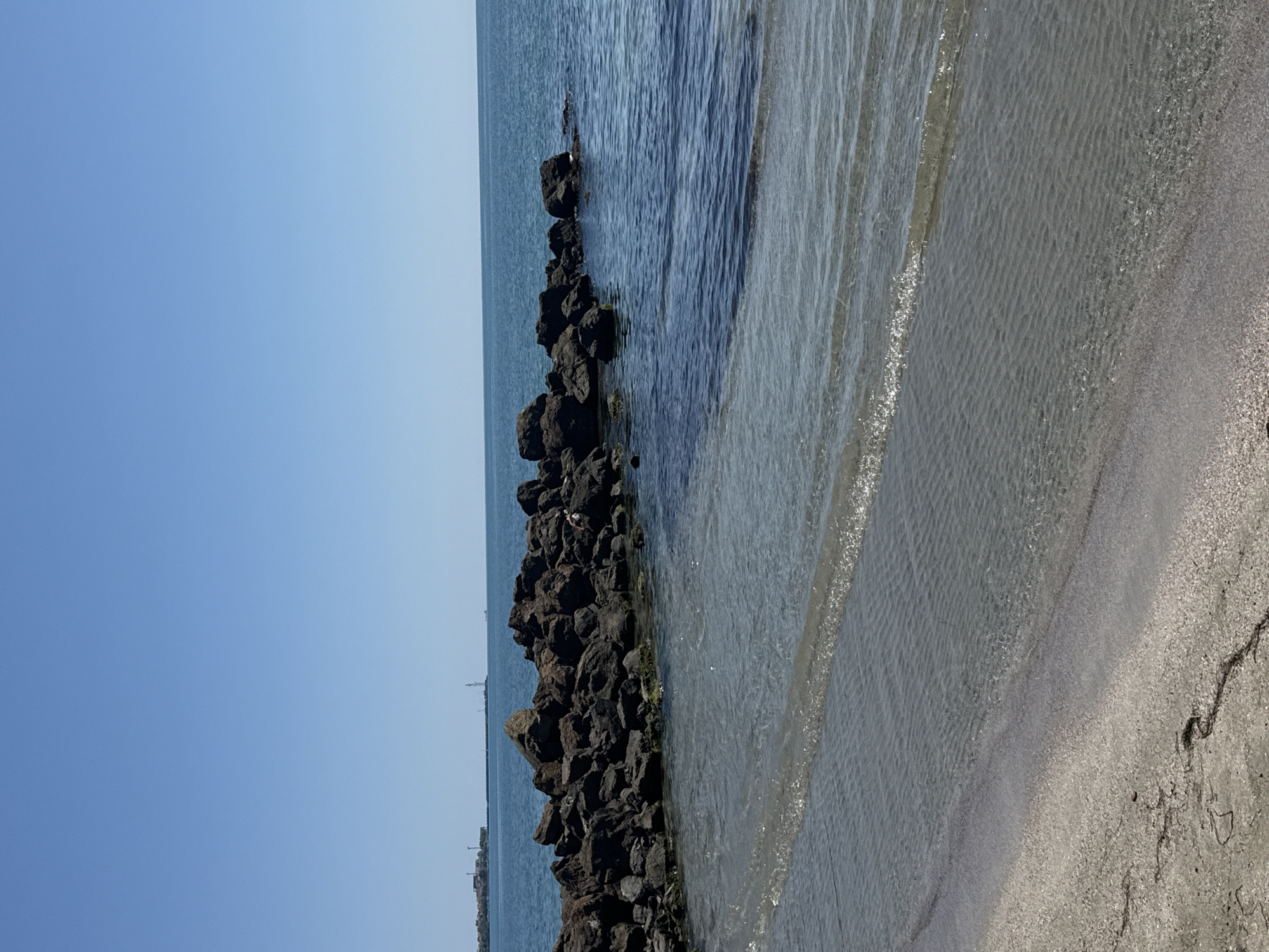 The image depicts a coastal scene in the city of Pomorie, Bulgaria, characterized by a calm sea and a rocky breakwater. The water is clear, with gentle waves lapping at the sandy shore. The breakwater, composed of dark, rugged rocks, extends into the sea, creating a sheltered area. In the distance, industrial structures and a lighthouse are visible on the horizon, suggesting the presence of a port or industrial zone. The sky is clear and blue, indicating fair weather. A single person is observed lying on the rocks of the breakwater. The overall impression is one of a serene and somewhat industrial coastal environment.