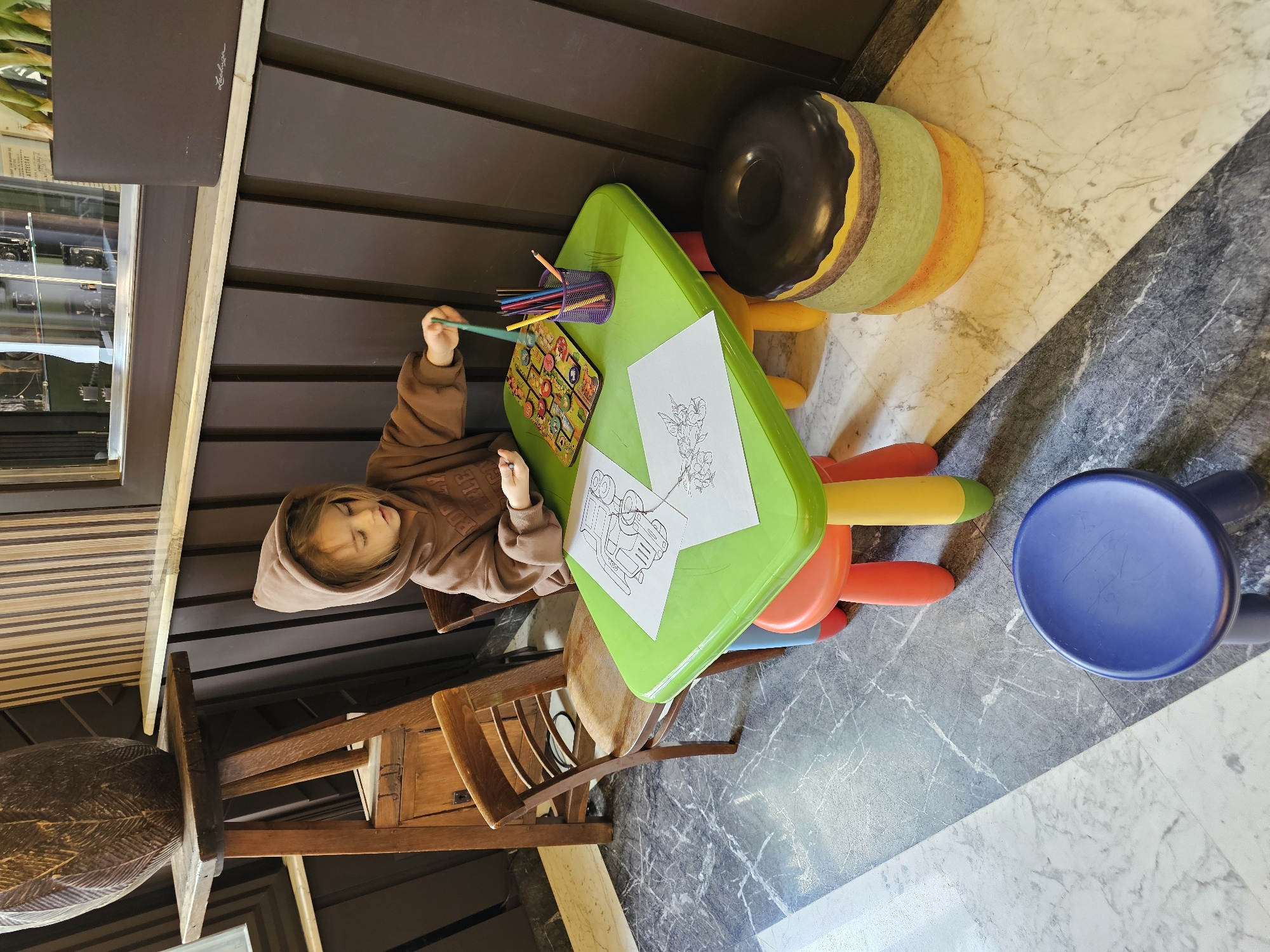 A child wearing a brown hooded sweatshirt is sitting at a green table with a wooden chair, coloring on a sheet of paper with colored pencils. The table also has a pencil holder and a game. Next to the table is a donut-shaped ottoman and a blue stool. The scene appears to be indoors, likely in a play area or waiting room. The floor is a grey-and-white marbled tile. Based on the provided information, this image was captured in Архангельское (красногорский Район), Russia.