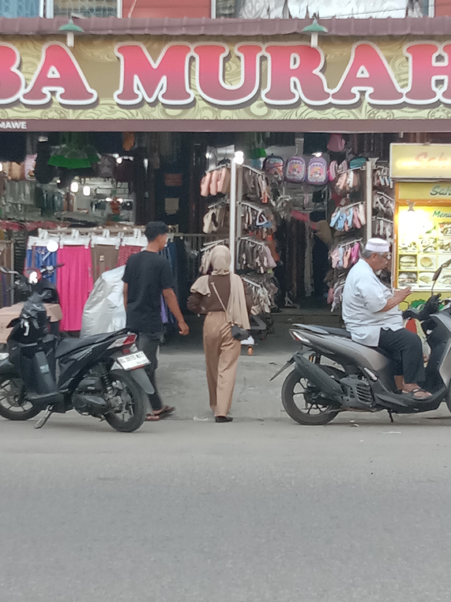 The image depicts a street scene in Lhokseumawe, Indonesia. A shop with a sign reading "BA MURAH" (translated as "Cheap Goods") is visible in the background. In front of the shop, a man is sitting on a moped looking at a cell phone. A woman in a tan hijab and pants, carrying a purse, walks with another man towards the shop. Two black mopeds are parked on the street.