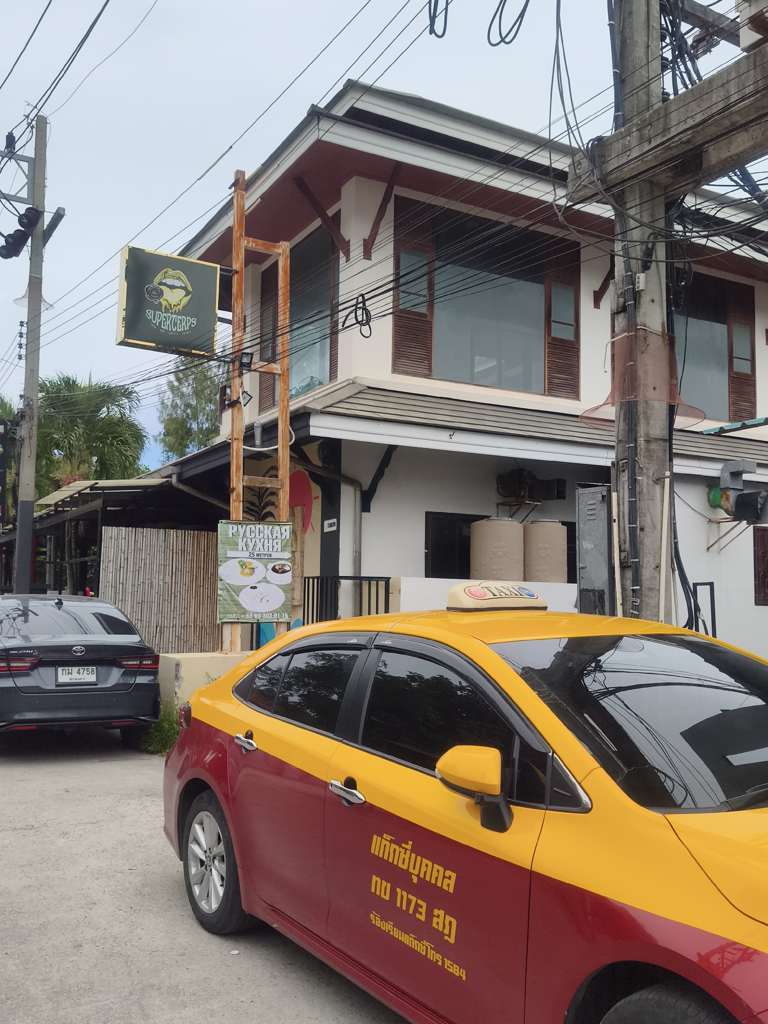 A two-story building with white and dark red accents is visible on a street in Baan Lamai, Thailand. A sign for "SUPERCERPS" hangs from a metal pole. A dark gray Toyota sedan with license plate "กท 4758" is parked on the left. A yellow and red taxi is parked to the right. A sign in Russian advertising "Русская Кухня" (Russian Cuisine) with a phone number is also visible on the side of the building. The sky is overcast.