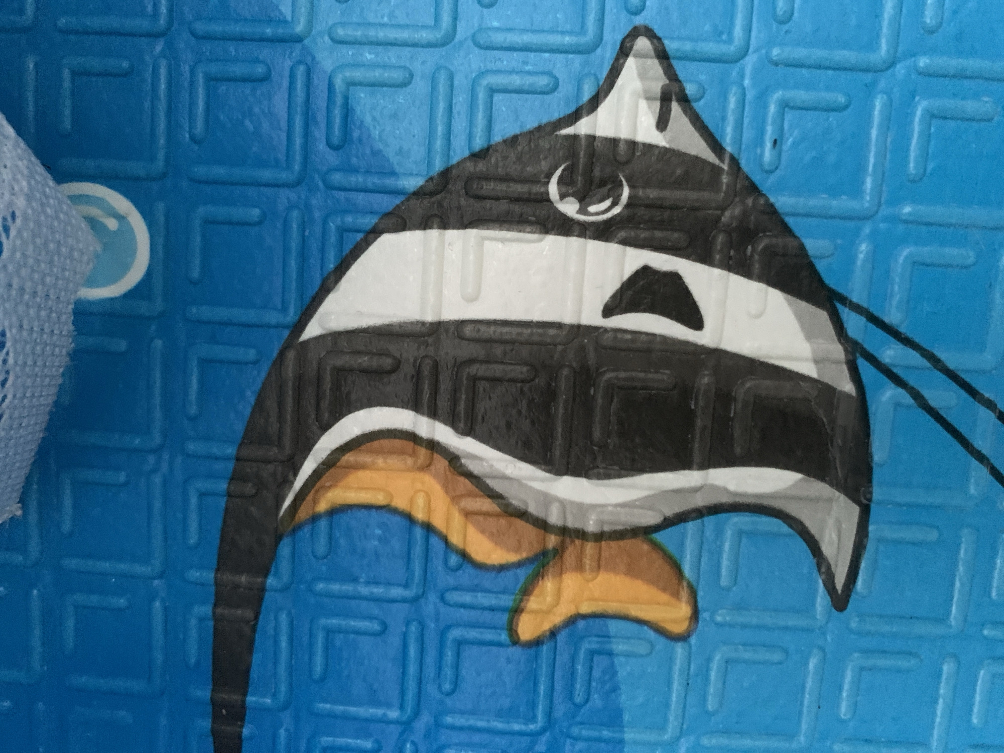 The image is a close-up of a cartoonish depiction of a Moorish idol fish. The fish has black, white, and gray vertical stripes, with an orange dorsal fin and tail fin. It is depicted against a bright blue background with a textured, grid-like pattern. A small portion of a white, textured fabric is visible at the top of the image, obscuring a small part of the fish's head. There is no discernible location in Stavropol, Russia, as the image is a graphic illustration and not a photograph of a real-world scene.