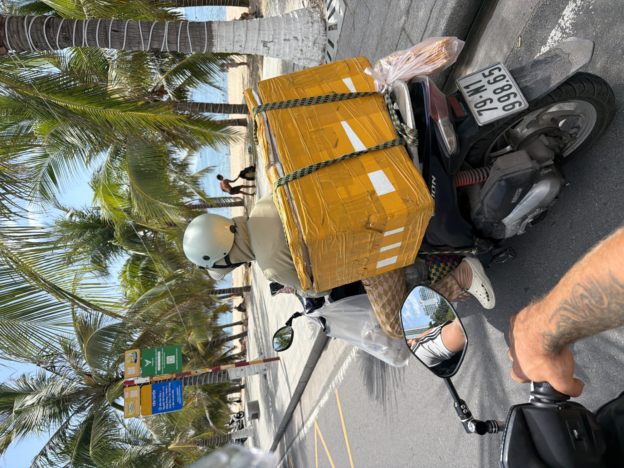 This photograph captures a vibrant street scene in Nha Trang Ward, Vietnam, from the perspective of someone riding a motorcycle. The setting is a sunny coastal area, indicated by the clear blue sky, numerous palm trees lining the road, and the visible ocean and sandy beach in the background.
The central subject is another individual riding a scooter, wearing a white helmet and light-colored clothing. This person is heavily laden with two large, yellow-wrapped packages securely strapped to the rear of their scooter with a multi-colored bungee cord. A plastic bag, possibly containing bread or snacks, is also attached near the packages. In the background, on the beach, a shirtless person can be seen near the water, enjoying the clear weather.
Notable details include the lush palm trees, some with white bands wrapped around their trunks, and the asphalt road with yellow markings. Road signs attached to a pole are visible on the left, one green with a QR code and a logo resembling VinFast, and a blue one with white Vietnamese text. The blue sign reads "TÊN TUYỂN" (Name of Route/Line) and lists street names such as "Nguyễn Tài Thánh," "Trần Phú," and "Tân Thái Tông."
The motorcycle carrying the packages is a Honda Vision, as indicated by the "HONDA" text near the license plate and "VISION" on its side. The license plate clearly displays "79-N1 998.65." In the foreground, the tattooed arm of the person taking the photo is visible, along with their motorcycle's rearview mirror, which reflects tall, modern buildings and a glimpse of the photographer's legs in striped shorts. The overall impression is one of a busy, sunny day in a Vietnamese coastal city.