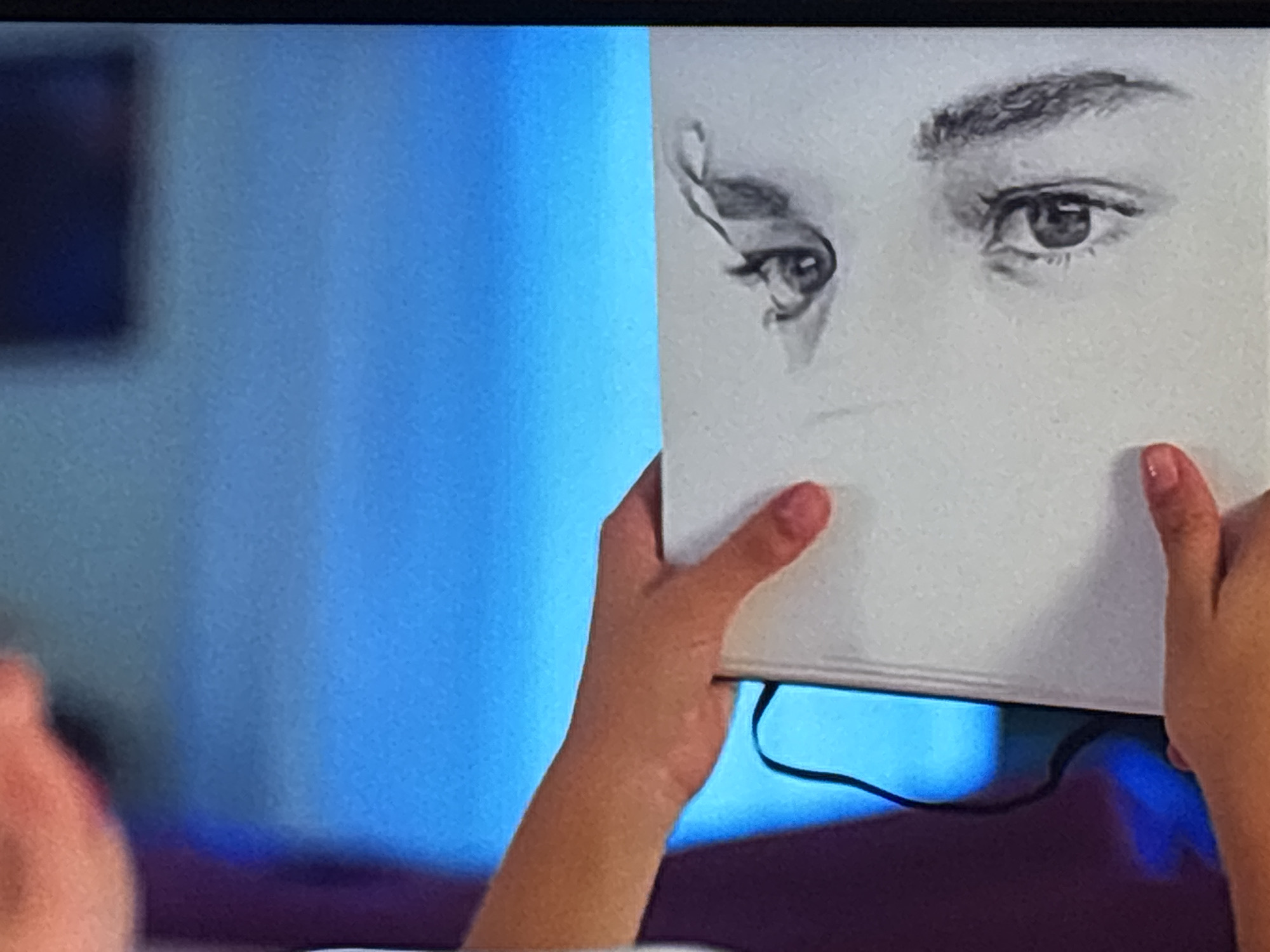 A person is holding up a sketchbook with a pencil drawing of a pair of eyes, obscuring part of their face. The drawing shows detailed eyes and eyebrows. The person's hands and arms are visible, holding the sketchbook. A blurred blue background is present. No information in the image indicates the location is Sofia, Bulgaria.