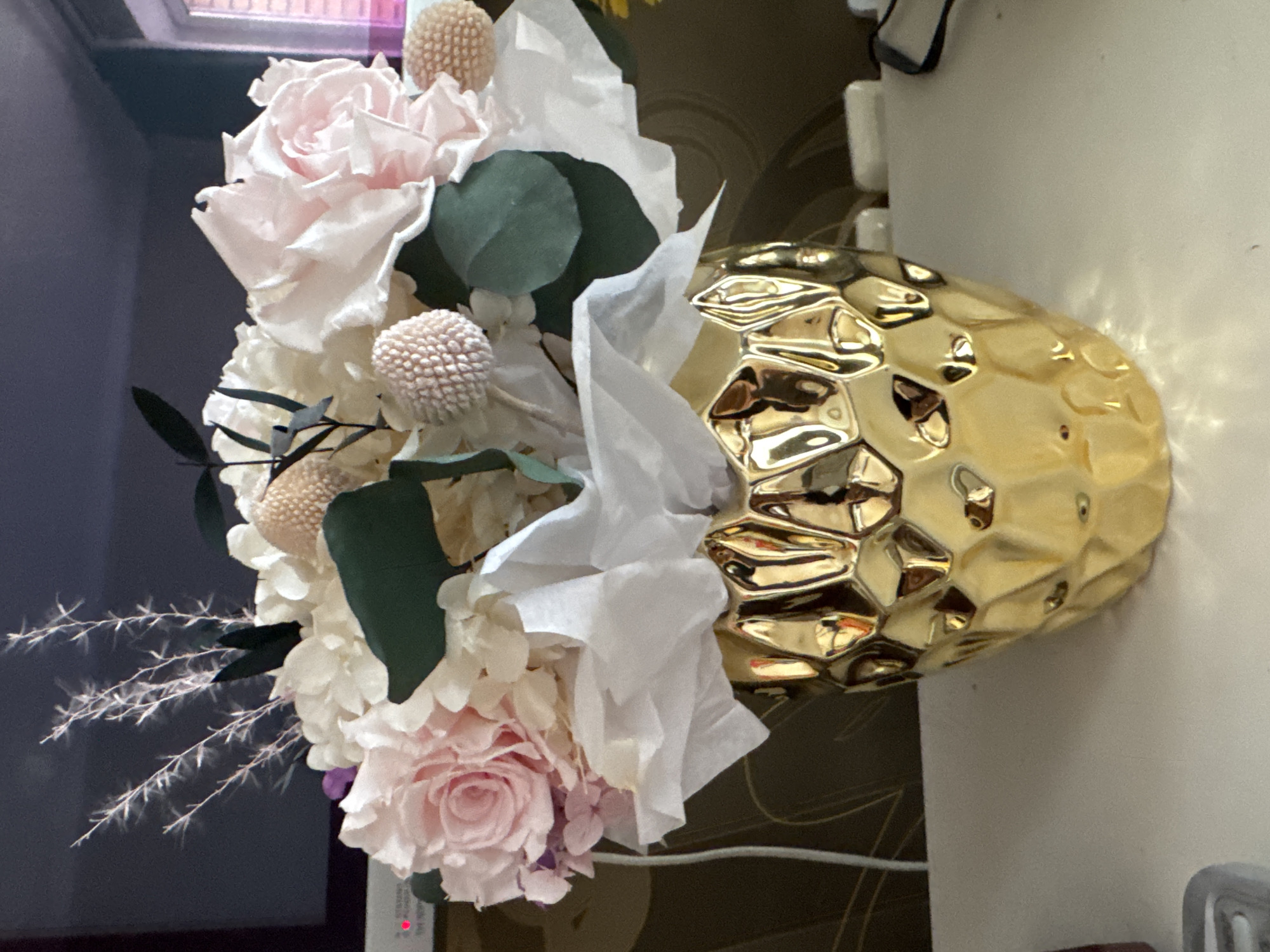 A bouquet of light pink and white flowers is displayed in a textured gold vase. The flowers include roses, hydrangeas, and other filler flowers, arranged with green leaves and white tissue paper. The vase has a geometric, hammered design. The background is indistinct, suggesting an indoor setting.