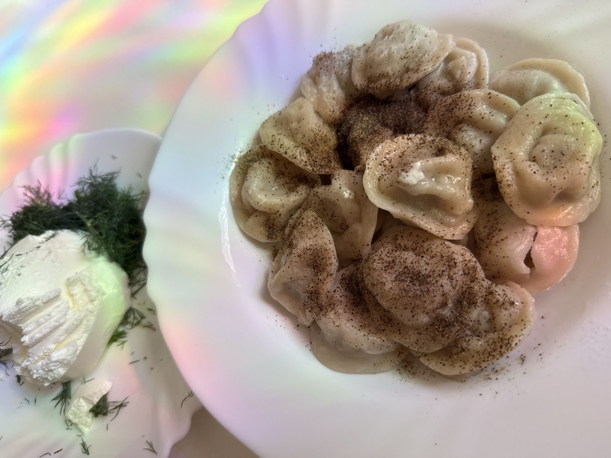 Two white plates with scalloped edges are visible in the image. The plate in the foreground contains a pile of pelmeni, a type of dumpling, sprinkled with black pepper. The pelmeni are light-colored and appear to be boiled. The plate in the background holds a dollop of white cream cheese or sour cream, garnished with fresh dill sprigs. Both plates are positioned on a surface that reflects a rainbow of colors, suggesting diffused lighting or a holographic effect. The background is out of focus, emphasizing the food. The scene is static and appears to be a close-up shot of a meal.