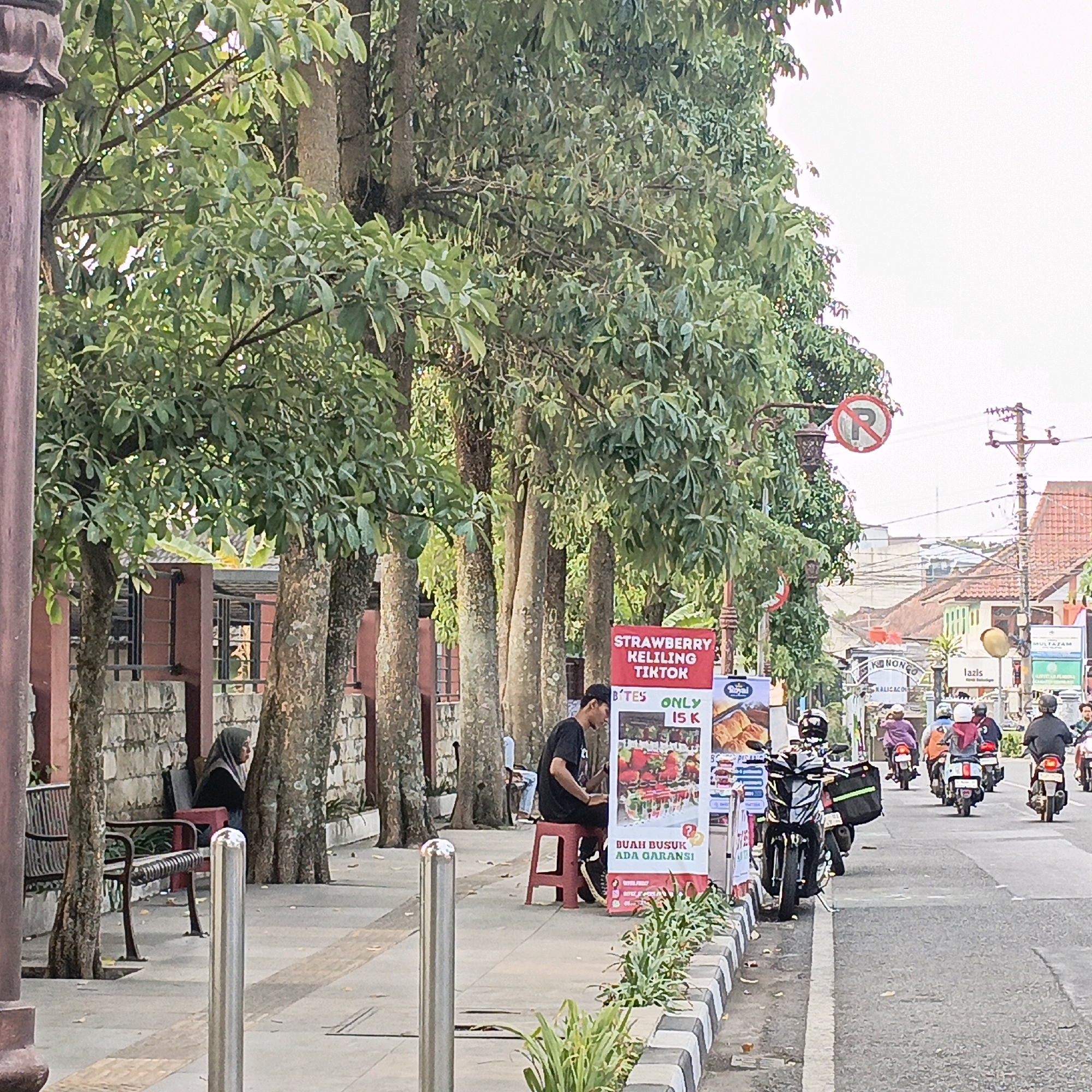 This is a street scene in Salatiga, Indonesia. The foreground shows a sidewalk lined with trees and a metal bench. A person in a hijab is seated on the bench. A young man in a black t-shirt and cap is seated on a red stool next to a sign advertising "STRAWBERRY KELILING TIKTOK" and "BUAH BUSUK ADA GARANSI" (rotten fruit guaranteed). Several motorcycles are parked on the side of the road. In the background, there are buildings with red roofs and a busy street with people on motorcycles. A "no parking" sign is visible on a lamppost. The overall impression is a lively street scene in a tropical climate.