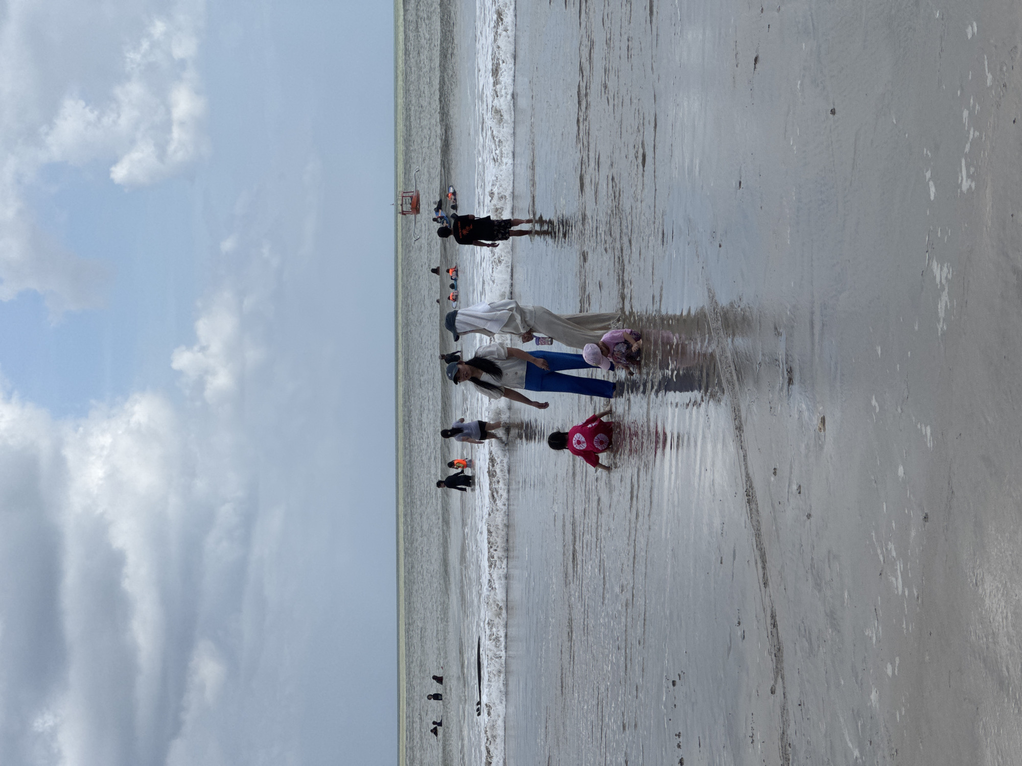 The image depicts a beach scene in Kuta Selatan, Indonesia, under a cloudy sky. Several people are present in the shallow water and on the wet sand. In the foreground, two young children are playing in the water, one in a pink garment and another in a pink hat. To their right, two women are standing; one in a white top and blue pants, and the other in a light-colored headscarf and matching pants. Further back, more individuals are visible in the ocean, some appearing to be swimming or wading. In the distance, a small orange boat with a canopy is anchored in the water, and other people are seen on surfboards. The waves are moderate, and the water is calm around the people. The sand appears wet and reflective due to the receding tide.