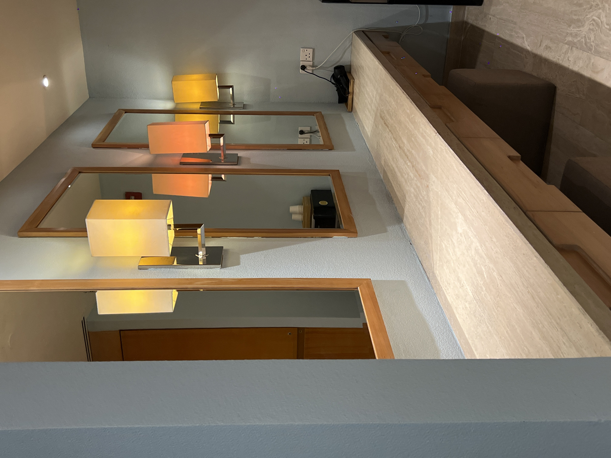 The image depicts a hotel vanity area, likely in a room in Nha Trang Ward, Vietnam. The scene features a long, light-colored countertop with wooden drawers underneath. Above the countertop, several wall-mounted lamps with rectangular shades cast warm light. Three framed mirrors are positioned above the lamps, reflecting the lamps and the surrounding wall. On the countertop, a black hairdryer is visible on a wooden tray near electrical outlets. To the left, a portion of a wooden cabinet is reflected in the leftmost mirror. The walls are painted a light blue, and the floor is tiled with a marble-like pattern. The overall impression is one of a well-lit, functional, and modern vanity space.