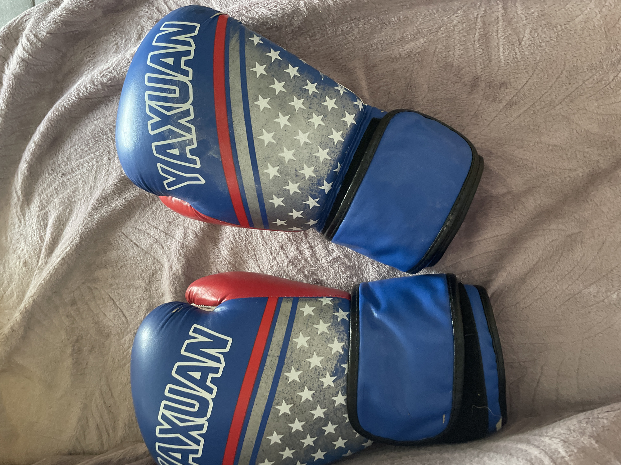 This media file displays a pair of blue boxing gloves with a red thumb and red and white stars on a gray background, superimposed over a red, white, and blue stripe. The word "YAXUAN" is printed in white capital letters on each glove. The gloves are lying on a pink, textured blanket. The lighting suggests an indoor setting. The image is a still shot, with no activity or event taking place. The gloves appear to be new or well-maintained, with no visible signs of wear or damage. The focus is on the gloves themselves, with the blanket serving as a neutral backdrop. The overall impression is one of readiness or preparation for a sporting activity.
