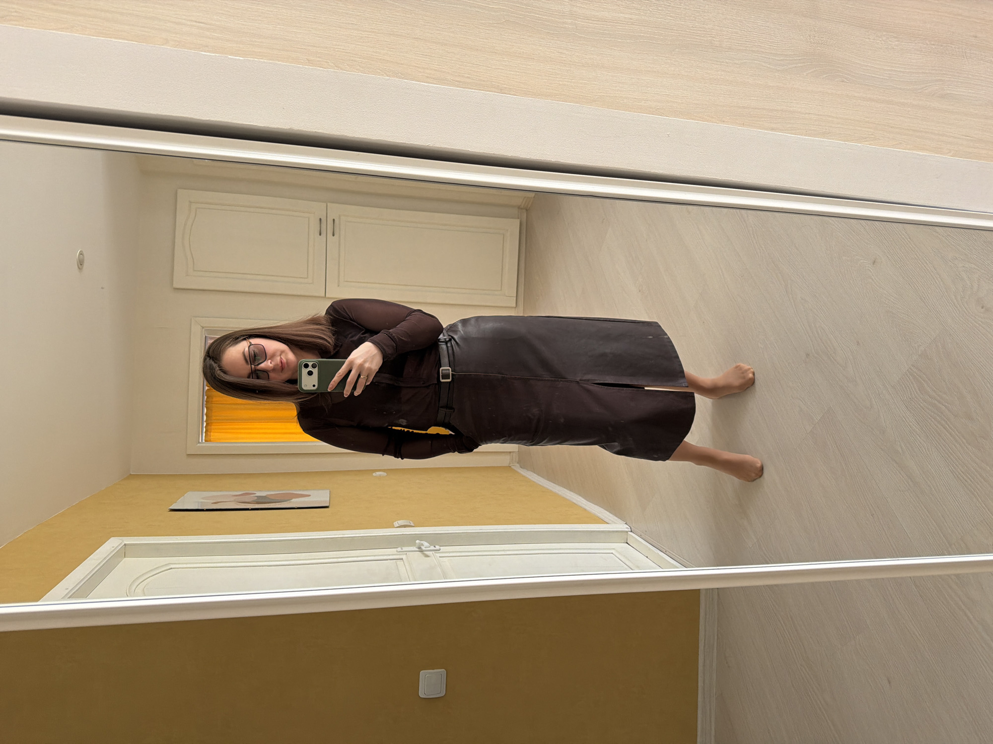 A medium-shot of a woman reflected in a mirror, seemingly indoors. The woman is wearing glasses, a long-sleeved brown top, and a dark brown leather skirt with a belt. She is holding a green smartphone, presumably taking the photograph.
The room's walls appear to be painted in shades of yellow and white. There's a door with white trim visible in the background, as well as some closed white cabinets. The floor is light-colored wood. The setting does not obviously appear to be within the city of Izhevsk, Russia.