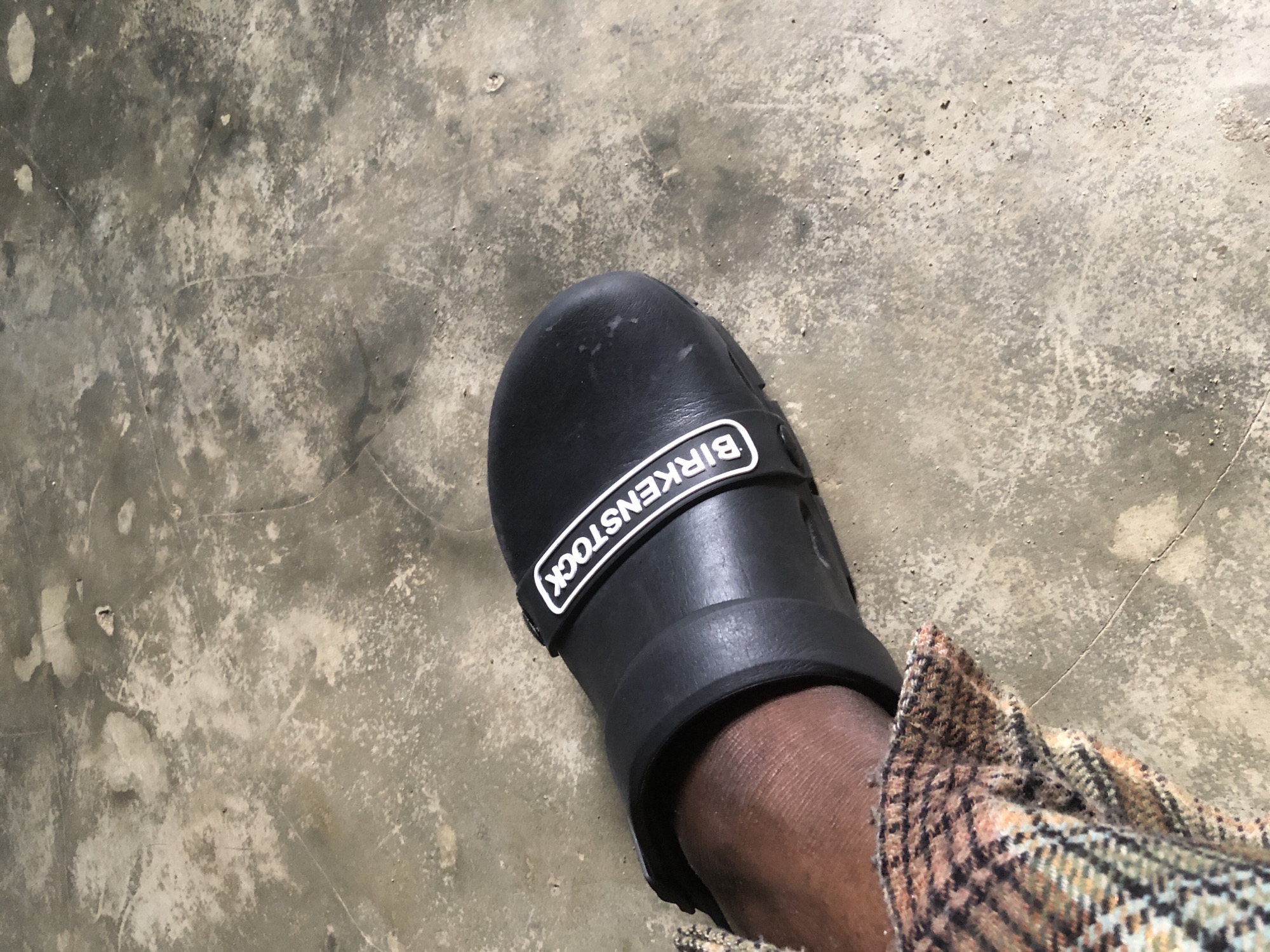 A close-up, low-angle shot shows a black Birkenstock clog on a person's foot, positioned on a concrete floor. The clog has the "BIRKENSTOCK" logo in white letters on a black strap across the top. The person's ankle and lower leg are visible, covered by a plaid fabric in shades of brown, orange, and black. The concrete floor is textured and shows signs of wear, with variations in color and some small debris. The image is taken indoors, likely in an unadorned space. The scene appears to be in Ado, Nigeria, where such practical footwear and simple flooring are common.