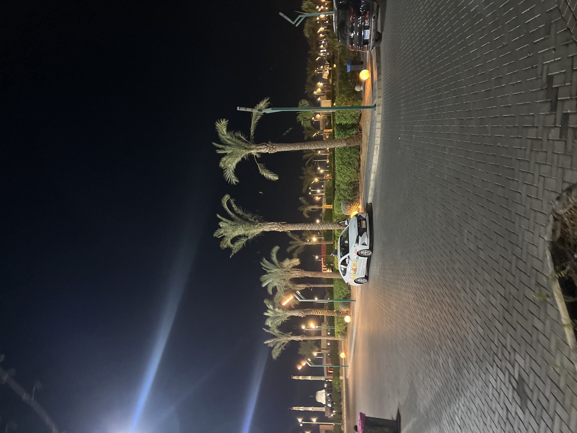 The image depicts an outdoor nighttime scene in Sharm El Sheikh, Egypt. The foreground is paved with dark gray interlocking bricks, arranged in a herringbone pattern. To the left, a dark gray trash bin with a pink lid is partially visible.
In the middle ground, a white sedan, possibly a taxi, is parked on the right side of a paved roadway. The car has colorful stickers on its side. Behind the car is a neatly trimmed green hedge. Several tall palm trees with lush green fronds are spaced along the hedge, their trunks illuminated by overhead lights. Some of these lights are mounted on modern, thin poles with a unique angular design.
Further in the background, a row of similar palm trees and hedges continues. Beyond that, illuminated structures are visible, including what appear to be minarets of a mosque or religious building on the far left, along with other buildings and illuminated trees and decorative spherical lights on the ground. The sky is a deep, dark blue-black, indicating nighttime. A bright light source, possibly a street lamp or vehicle headlight, creates a strong glare and lens flare in the upper left portion of the image. A dark-colored SUV is parked to the right of the white sedan. The overall impression is of a well-lit, modern urban or resort area at night.