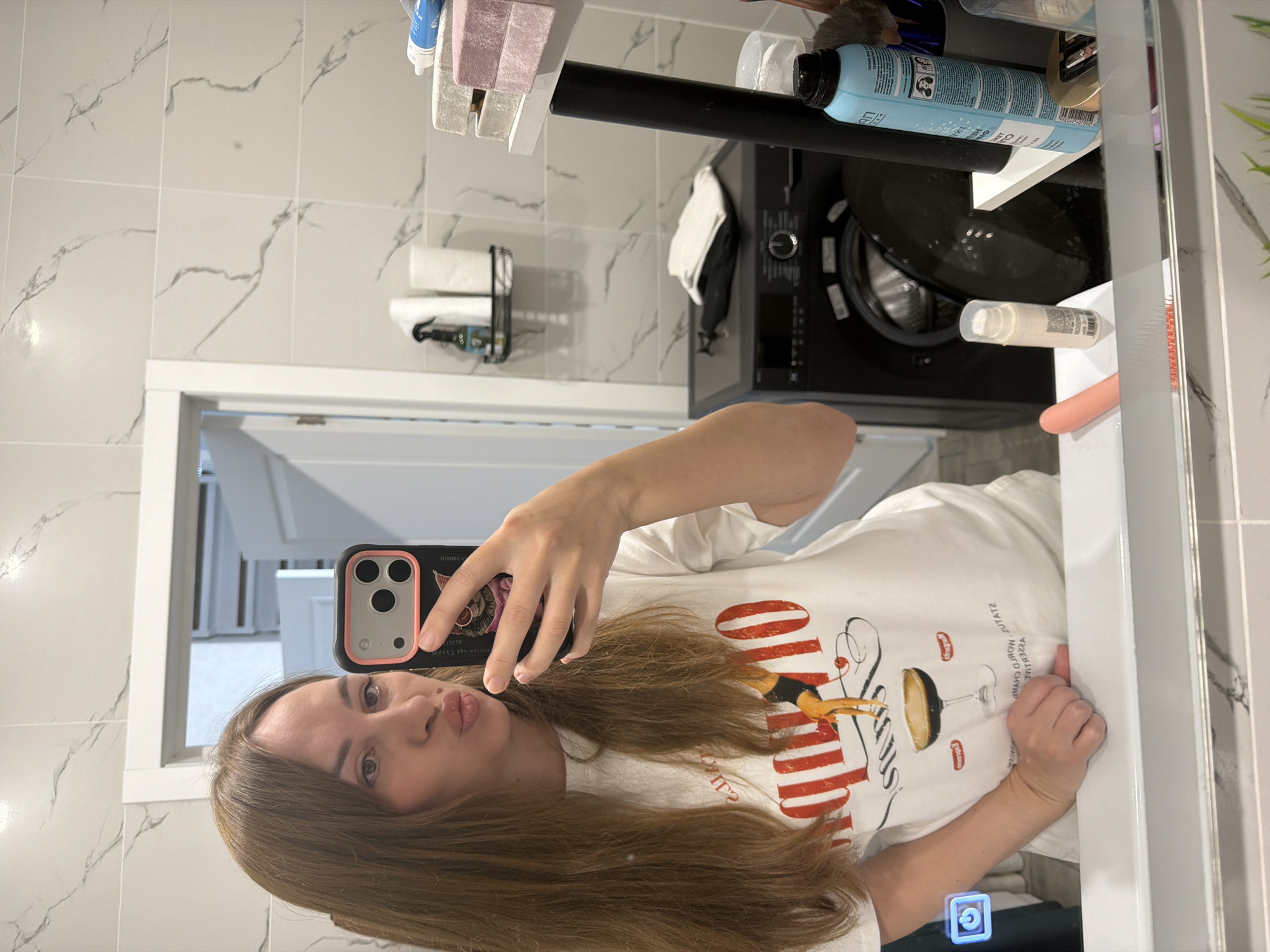 A woman with shoulder-length, light brown hair is taking a mirror selfie in a bathroom. She is wearing a white t-shirt with a graphic of a cocktail glass and the word "COCKTAIL" printed in red. The bathroom has white marble-patterned tiles on the walls and a white vanity with a large mirror. On a shelf above the vanity are toiletries, including spray bottles and paper towels. To the right of the woman, a black washing machine is visible. On the vanity counter, there is a pink object, a small white bottle with a pump, and a blue aerosol can. The mirror reflects the woman, the bathroom interior, and a doorway in the background. The setting is consistent with a residential bathroom.
