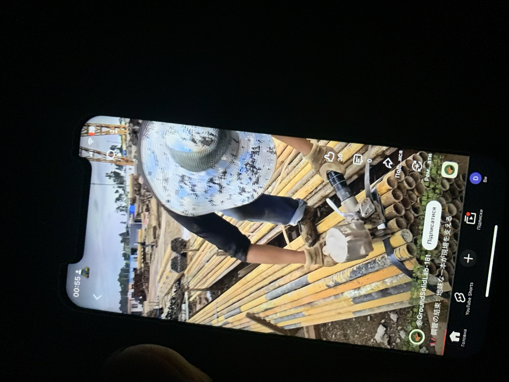The image displays a close-up view of a smartphone screen, held in landscape orientation. The screen shows a video playing, featuring a person wearing a camouflage-patterned hat and work clothes, engaged in an outdoor activity. The person is holding a tool and appears to be working with a large quantity of yellow pipes stacked around them. The background of the video shows an industrial or construction site with buildings and machinery.
On the phone screen, there are various UI elements, including a timestamp "00:55", a profile icon, a back arrow, and icons for likes (showing "30"), comments (showing "0"), and sharing. There are also text overlays in what appears to be Japanese characters, with the handle "@GroundSolidLab-18t" visible. At the bottom of the screen, a navigation bar for a video platform is displayed, with icons for "Home", "YouTube Shorts", a plus sign for creating content, and "Subscriptions" or "Notifications". The overall lighting suggests the photo was taken in a dimly lit environment, possibly at night or indoors, with the phone screen being the primary light source. There is no indication of the city of Zhmerynka, Ukraine, or any specific location within it.