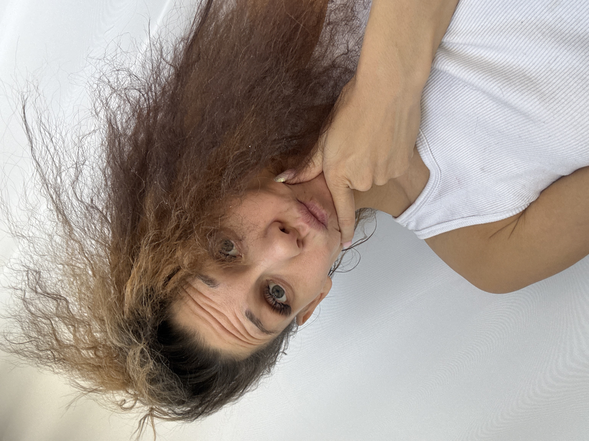 A woman is facing the camera with her head tilted to the left. Her hair is a mix of brown and lighter shades. She's wearing a white tank top. Her right hand is placed on her chin. The background is a plain white surface. The location is not discernible from the image.