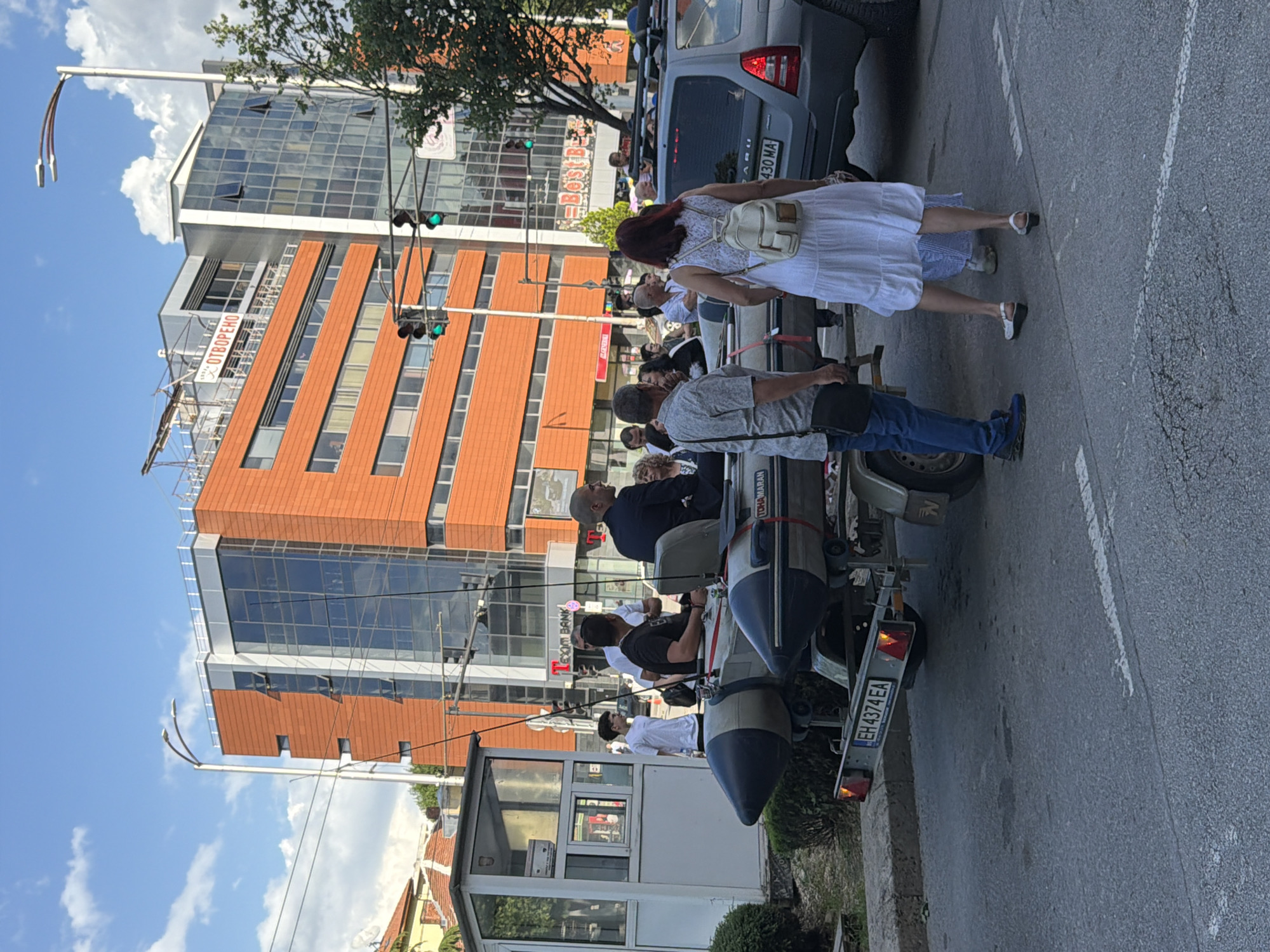 The image shows an outdoor scene in Pleven, Bulgaria. A group of people are being transported on a trailer pulled by a car. The trailer appears to be modified to carry passengers. The car is a gray Subaru. A woman in a white dress and a man in jeans and a gray shirt are walking next to the trailer. Buildings with modern architecture are in the background. A bus stop is visible. Traffic lights are also present. The license plate on the car is 430 MA. The license plate on the trailer is EH 4374 EA. The group is traveling on an asphalt road.