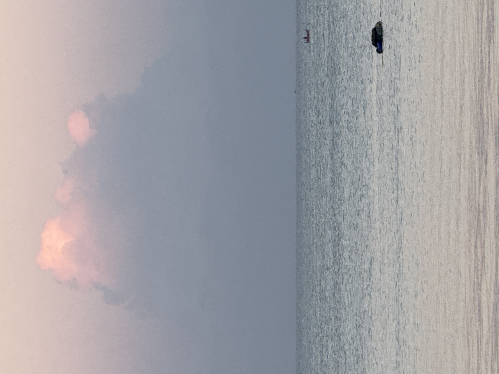 The image depicts a serene seascape with a muted color palette. A large, soft pink cloud dominates the upper portion of the sky, with a gradient of pale pink to light blue. Below the sky, the ocean stretches across the frame, its surface rippled and reflecting the soft light. The horizon line is clearly defined, separating the sky from the water.
In the distance, a small red and wooden boat with at least two people aboard is visible on the water. Further to the right, closer to the foreground, a dark object, possibly a buoy or a piece of debris with a blue netting attached, floats on the surface of the water. The overall impression is one of tranquility and solitude, with human presence minimal and integrated into the vastness of the ocean. The scene could be Kuta, Bali, Indonesia, known for its beautiful sunsets and coastal scenery.