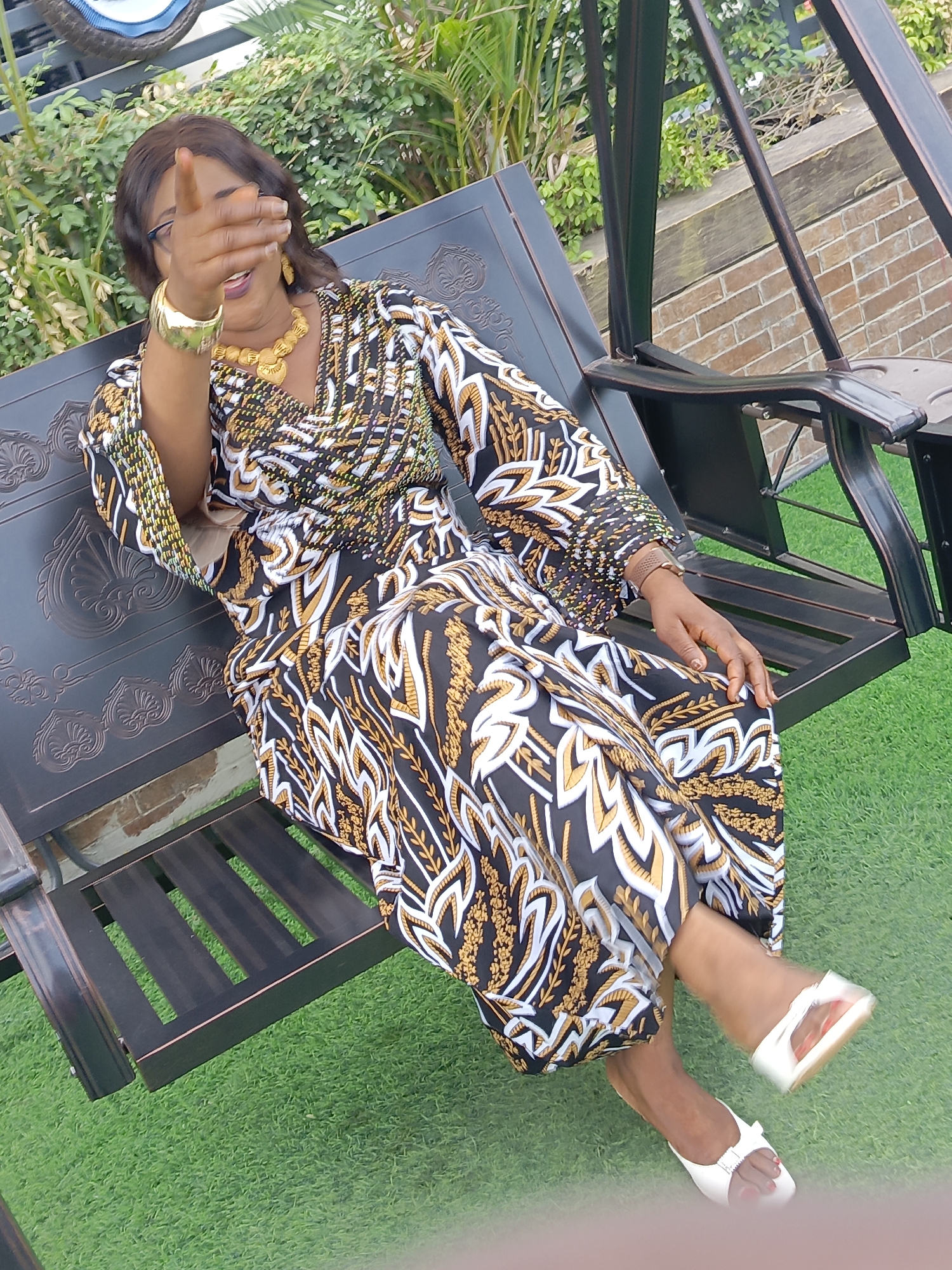 A woman is seated on a metal porch swing outdoors on an artificial green lawn. She is wearing a long-sleeved dress with a black, white, and gold patterned design and white sandals. Her left hand is raised, obscuring part of her face, and she is wearing gold jewelry on her neck and wrist. Green foliage is visible behind the swing, and a brick wall is present to the right. The woman is likely in Akwanga, Nigeria, though that information is not verifiable from the image.