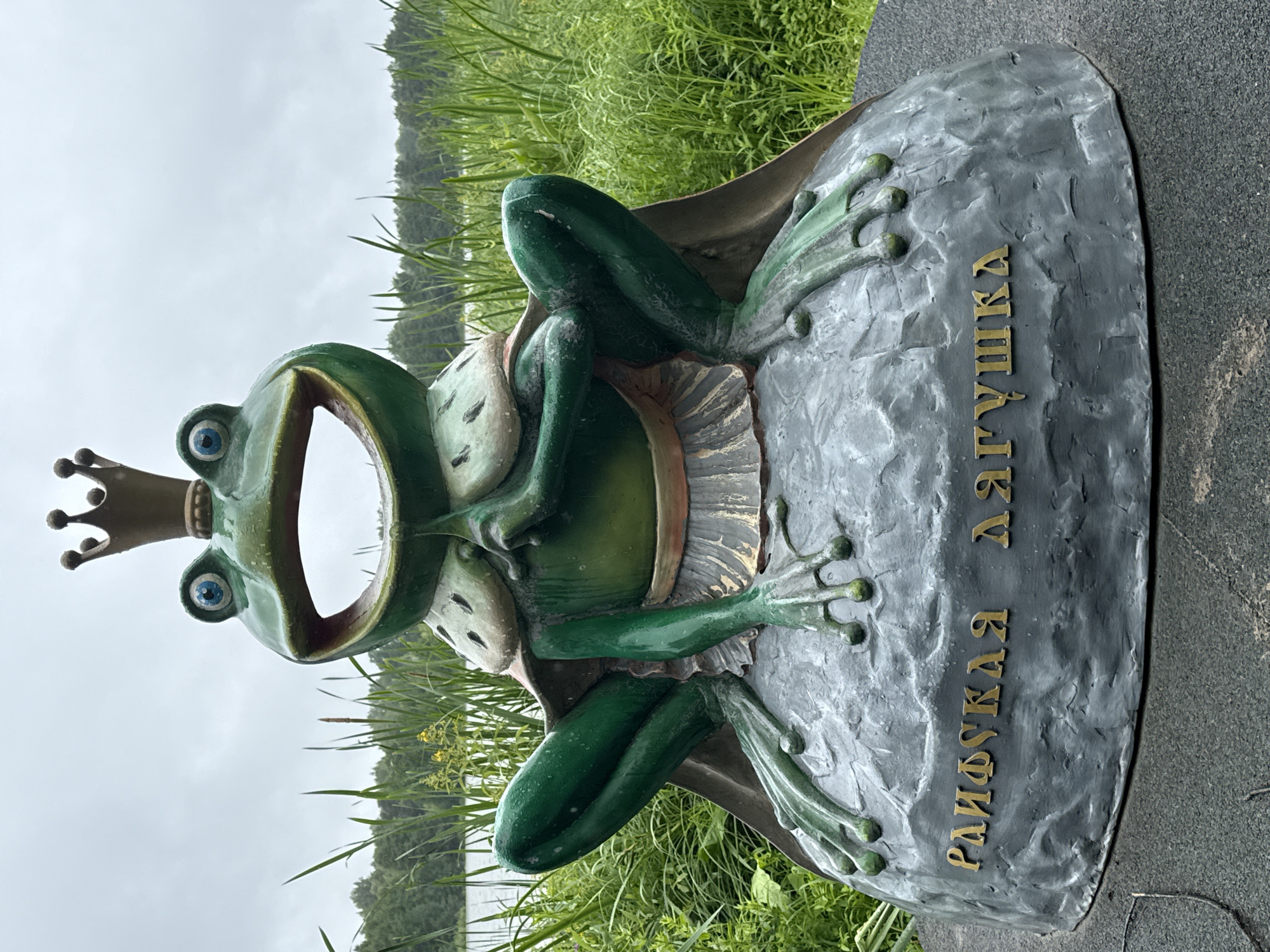 This is an upright, medium shot of a colorful statue of a frog wearing a crown, sitting on a rock. The frog is green with blue eyes and a wide, open mouth. It is wearing a beige cape and a white frilled skirt. The rock the frog is sitting on is grey and textured. There are gold-colored Cyrillic letters on the rock. To the left of the frog, there is a body of water with trees in the distance. The sky is overcast. The overall scene appears to be outdoors, possibly in a park or by a lake, given the presence of water, reeds, and grass. The setting is Местечко Раифа, Russia. The visible text on the rock reads "РАИФСКАЯ ЛЯГУШКА" which translates to "Raifa Frog".