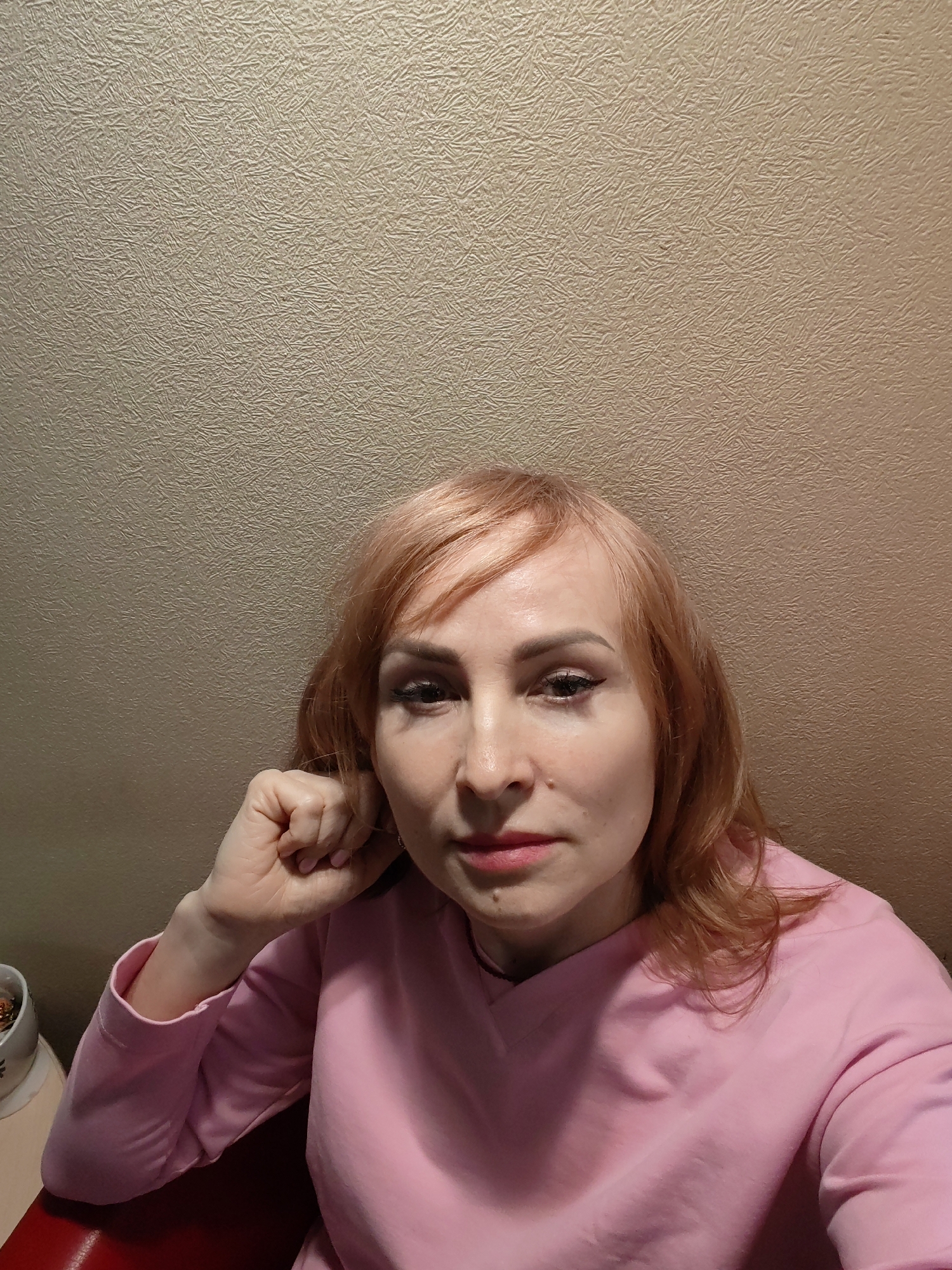 A woman with reddish-blonde hair and a pink long-sleeved shirt is looking at the camera with a neutral expression. Her left hand is touching her face, near her ear. She has a mole on her right cheek. The background is a textured beige wall. Part of a red chair and a small white container are visible in the bottom left corner of the frame. It is impossible to know whether the photograph was taken in Chaikovsky, Russia from the visual data provided.