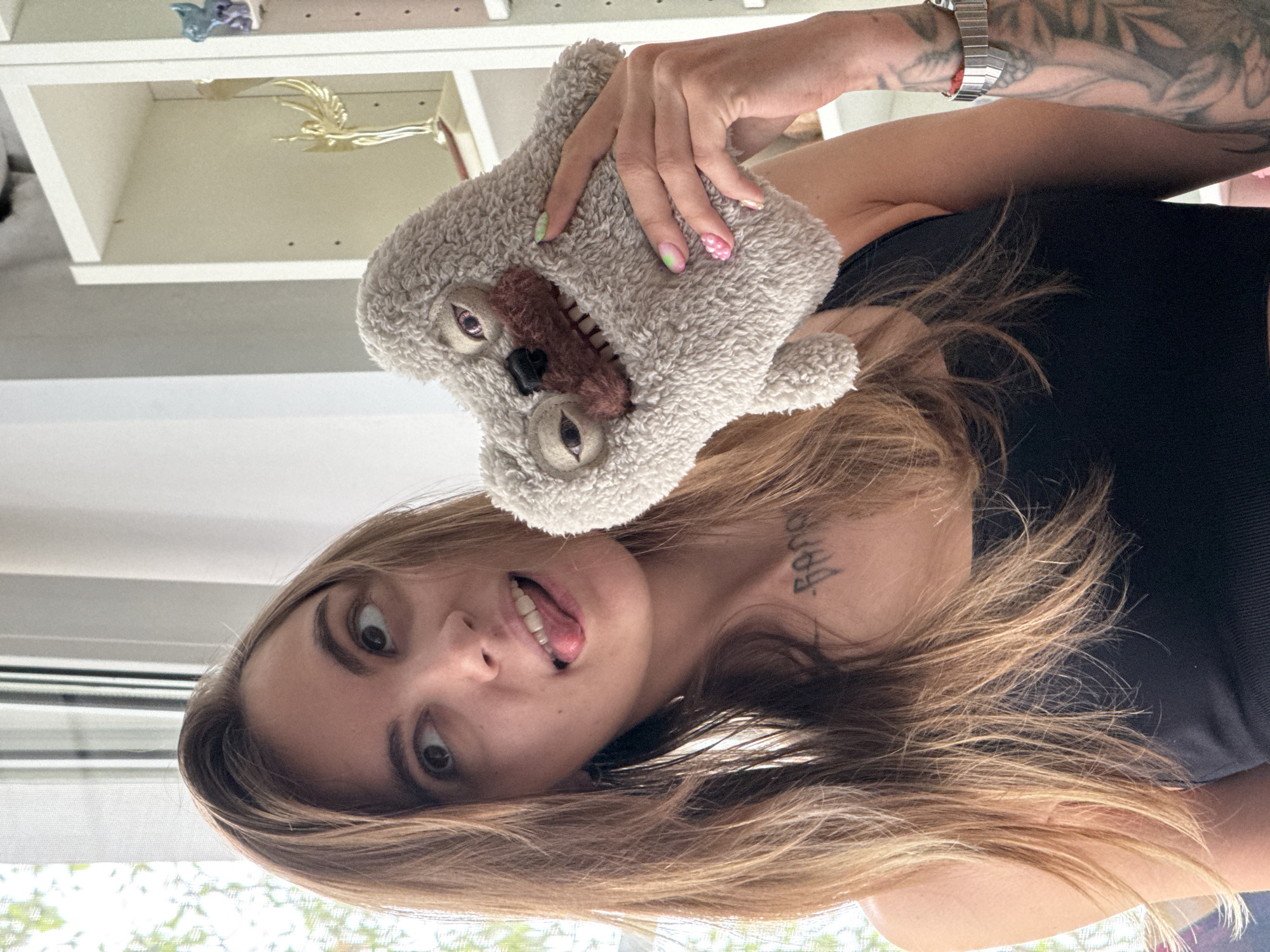 A young woman with long brown hair, wearing a black tank top, poses for a photo. She is holding a gray plush toy resembling a sloth, with its mouth open, and sticking her tongue out playfully. Her eyes are looking up and to the left. She has tattoos on her neck and arm. Behind her, a white shelf unit is visible, with various items on it, including a golden trophy. The scene appears to be indoors, with natural light coming from a window on the left. The exact location within Moscow, Russia is not determinable from the image.