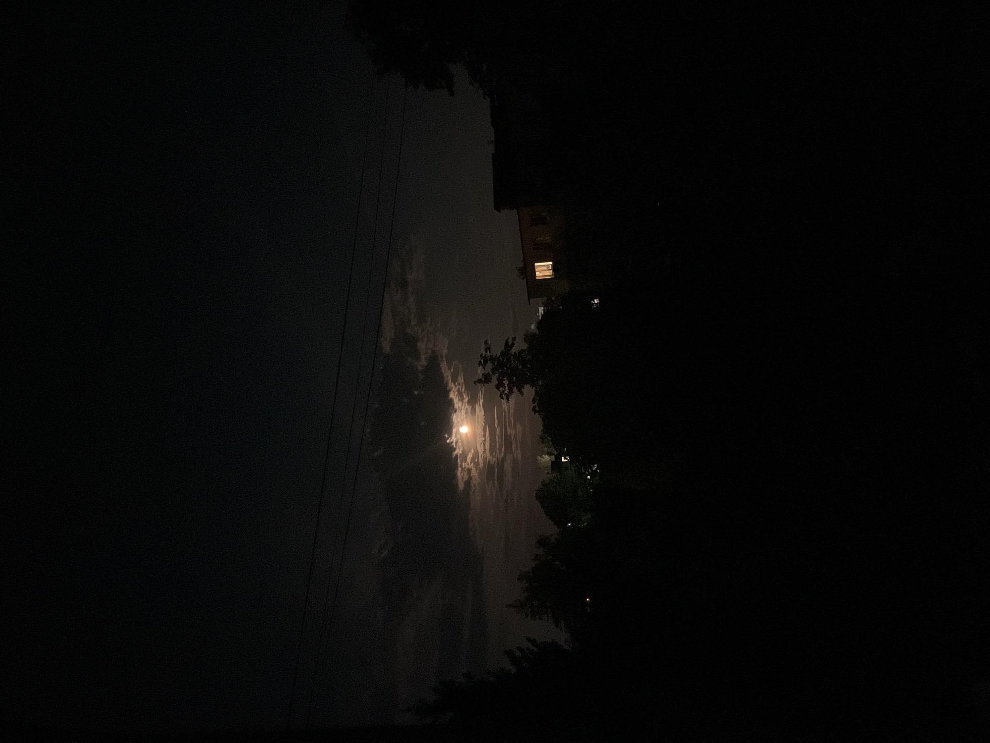 The image captures a nighttime scene in the city of Stari Sanzhary, Ukraine. The sky is dark with scattered clouds partially obscuring a bright, orange-hued moon. In the mid-ground, silhouetted trees and foliage create a dark, foreground element. A building with a single illuminated window is visible on the right side of the frame. Overhead, power lines are visible against the night sky. The overall impression is one of a quiet, possibly rural, residential area at night.