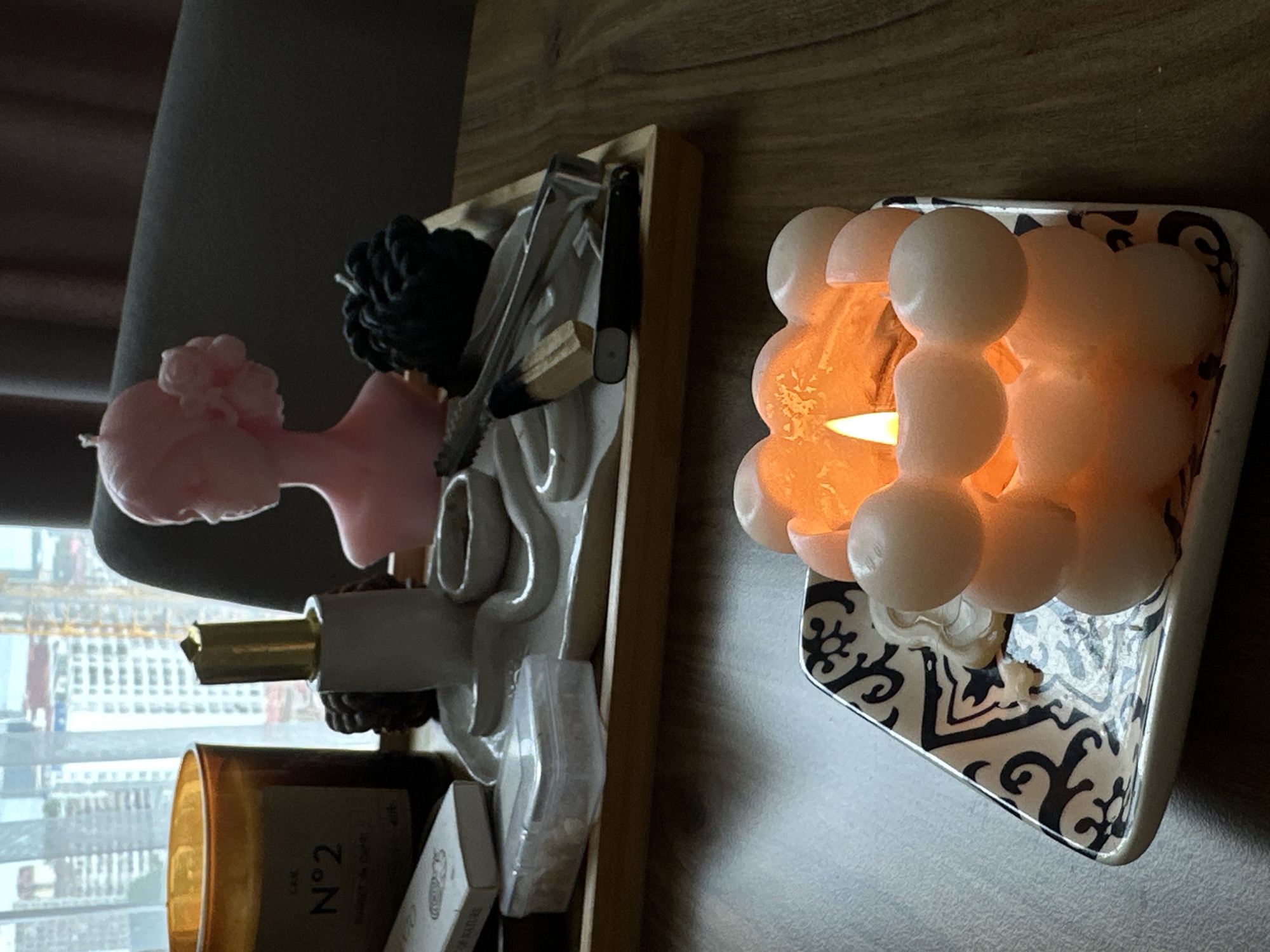 A dimly lit indoor scene focuses on two candles. In the foreground, a pink, bubble-shaped candle burns brightly on a decorative square plate with a black and white swirling pattern. In the background, a light pink bust candle sits on a wooden tray, alongside other items including a dark green knotted candle, tongs, a wooden stick, a black cylindrical object, a white dish with small indentations, and a clear plastic box. A large amber glass candle with the label "N°2" is also visible on the tray. Behind the tray, sheer white curtains reveal a cityscape with tall buildings and construction cranes, indicative of a city like Batumi, Georgia. The overall mood is serene and decorative.