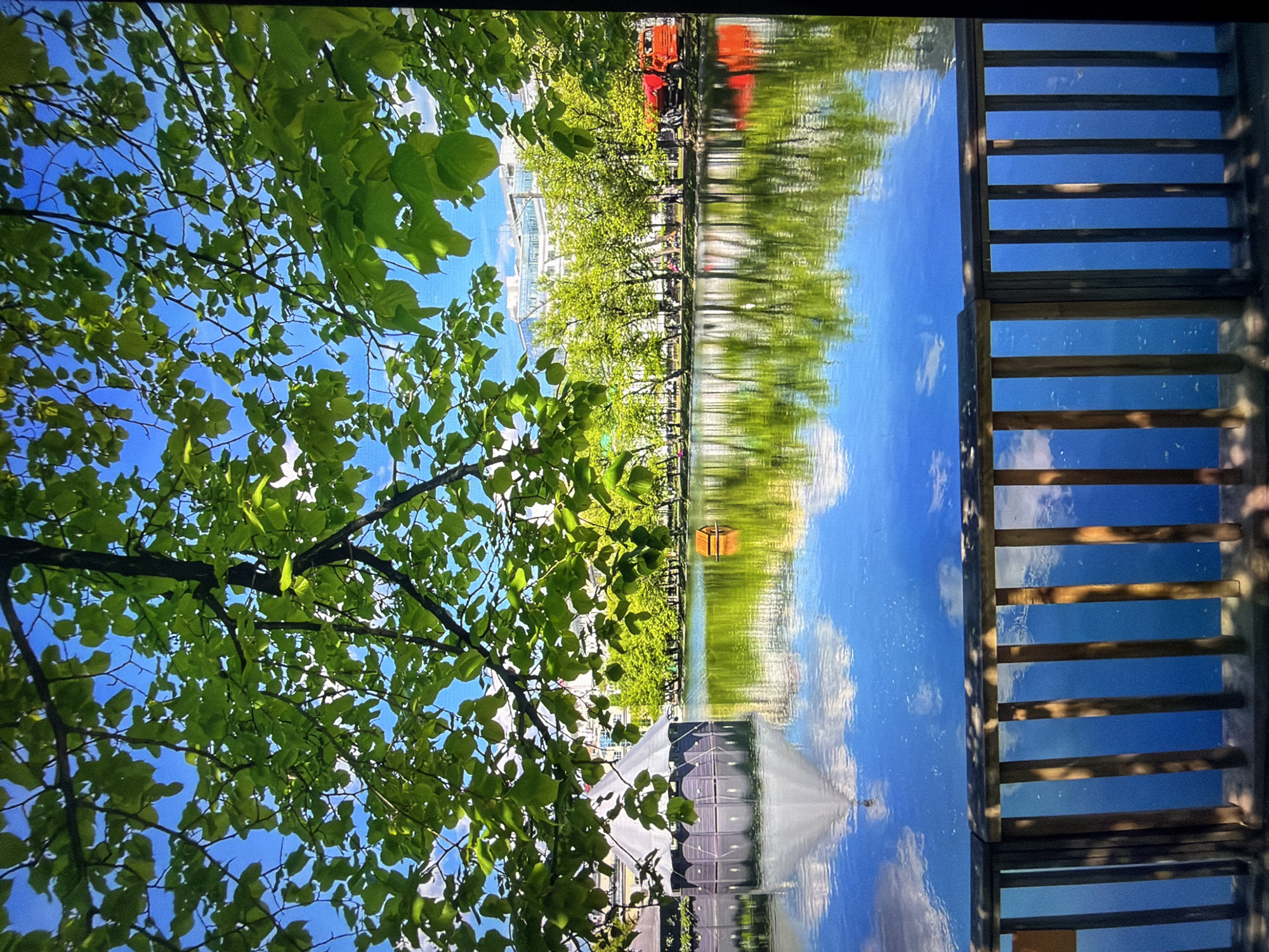 The image depicts a scenic view of a pond in the city of Kotelniki, Russia. In the foreground, a wooden railing with vertical slats runs across the bottom of the frame, casting shadows from the overhead foliage. Above the pond, lush green tree branches with vibrant leaves overhang, filtering the bright blue sky. The pond itself is a calm expanse of water, reflecting the surrounding environment with striking clarity.
On the left side of the pond, a unique, dark hexagonal structure with a light-colored, conical roof is partially submerged. This building appears to be a gazebo or a unique architectural feature. Further across the pond, a small, quaint wooden house, possibly a birdhouse or decorative element, floats on the water.
The far bank of the pond is lined with a dense row of vibrant green trees, their leaves illuminated by sunlight. Behind the trees, modern buildings with white facades and numerous windows are visible, suggesting an urban or semi-urban setting. A bright red truck is parked to the right of the trees, hinting at ongoing activity or maintenance in the area. Several indistinct figures can be seen strolling along the path behind the trees, adding a sense of human presence to the otherwise tranquil scene. The reflections of the trees, buildings, and sky in the water create a symmetrical and picturesque composition.