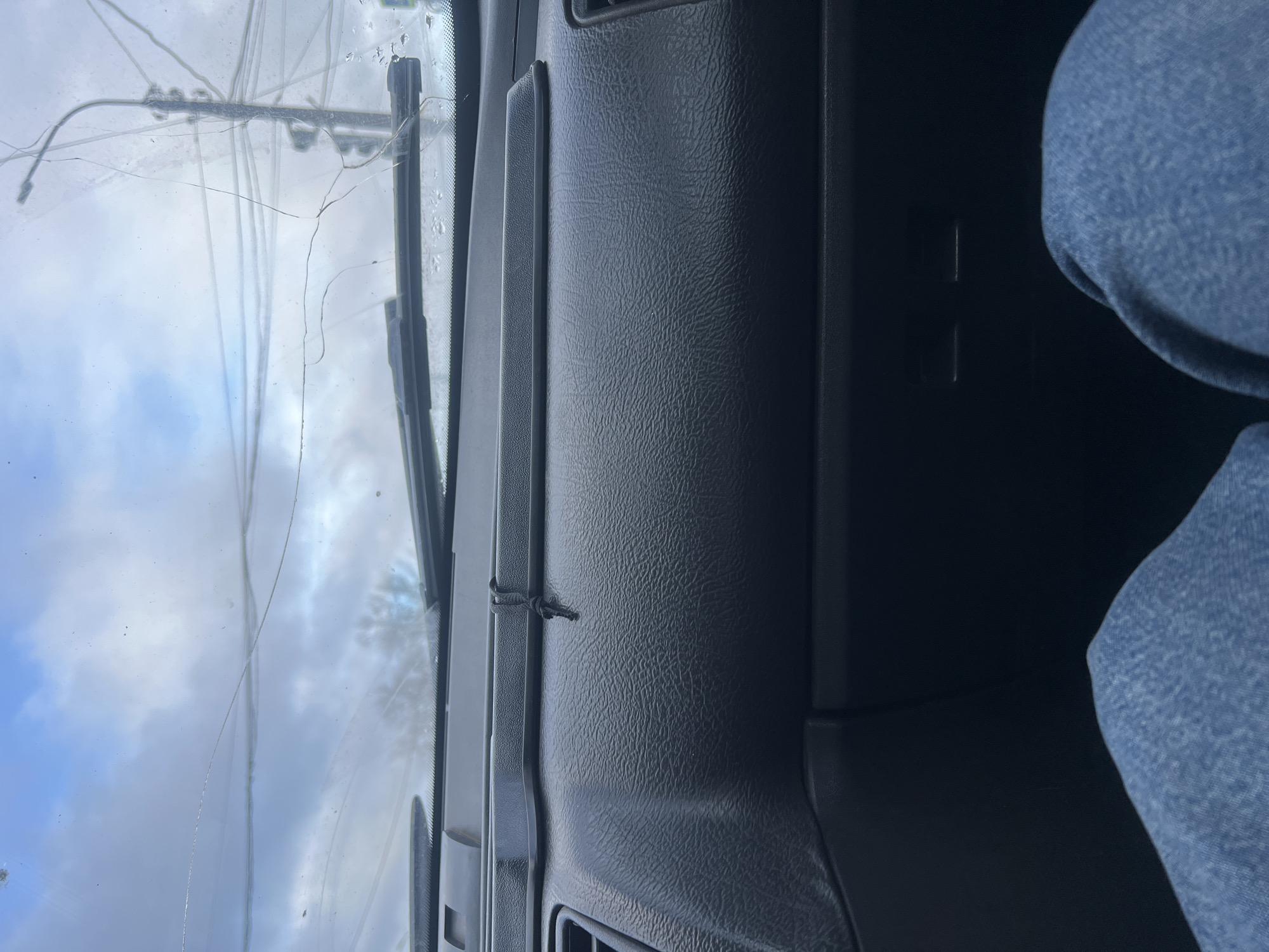 The image provides an interior view from a vehicle, looking towards the dashboard and through the windshield. On the right side, the knee and upper thigh of a person wearing blue denim pants are visible, indicating the presence of a passenger or driver.
The car's dashboard is made of dark, textured plastic, extending across the lower part of the frame. A small, black, knotted cord or tie is positioned on the dashboard near the base of the windshield.
The most prominent feature is the windshield, which is extensively cracked. Numerous long, linear cracks traverse the glass, varying in thickness and opacity, suggesting significant damage. Some dirt or water spots are also visible on the windshield surface.
Through the cracked windshield, a daytime sky with scattered clouds is visible, indicating either overcast or partly cloudy weather conditions. No specific activities are depicted beyond being inside a car. There is no discernible text visible in the image. The location context provided for the image is Chaikovsky, Russia.