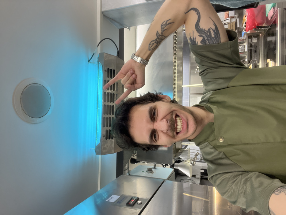A male individual is depicted in a commercial kitchen in Budva, Montenegro. He has dark, curly hair, a mustache, and a prominent smile, showing his teeth. He wears a green chef's jacket with a snap-button collar. His left arm is raised, forming a "rock on" hand gesture, with a silver-colored watch visible on his wrist. Tattoos are present on both arms; an abstract design is on his left forearm, and a dragon-like creature is on his right bicep. The background consists of stainless steel commercial kitchen equipment, including a unit marked "K-0005" on the left and various cooking surfaces. Overhead, a circular fixture and a rectangular insect trap emitting a blue light are visible, illuminating the ceiling.