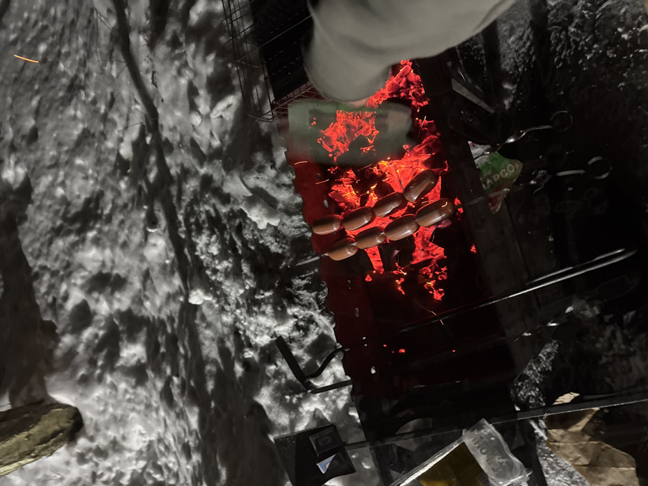 A bird's-eye view captures sausages grilling over an intense fire, the coals glowing a vibrant red. Snow surrounds the grill, creating a stark contrast between the cold exterior and the heated cooking area. Utensils and a package sit at the bottom. A partial view of a person's arm/sleeve.