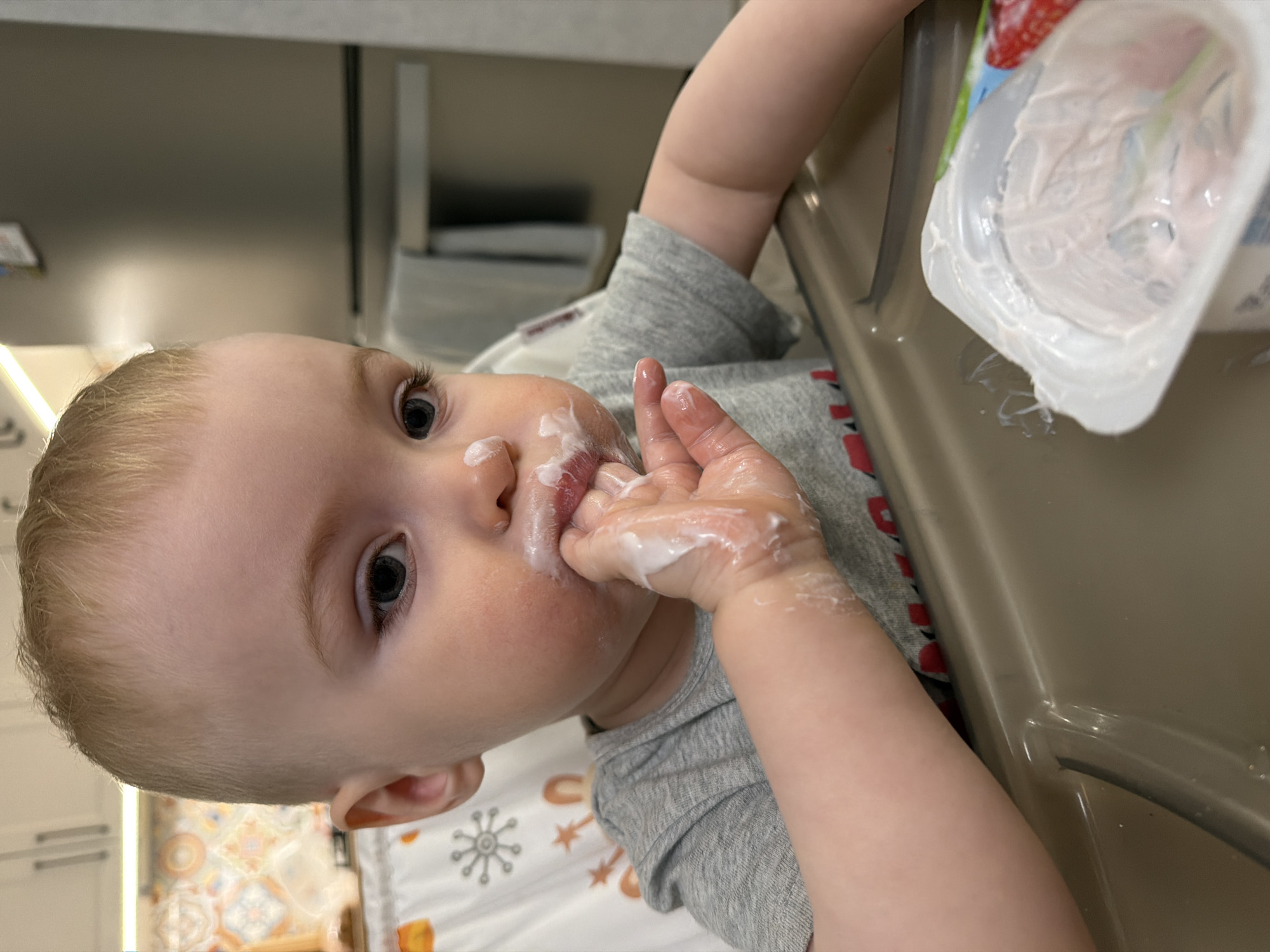 A young child is sitting in a high chair, eating yogurt. The child is fair-skinned with light blond hair, and their face and hand are smeared with yogurt. The child is wearing a gray long-sleeved shirt. A container of yogurt is visible on the high chair tray. The background shows a kitchen counter and a wall. The location and city cannot be determined from the image.