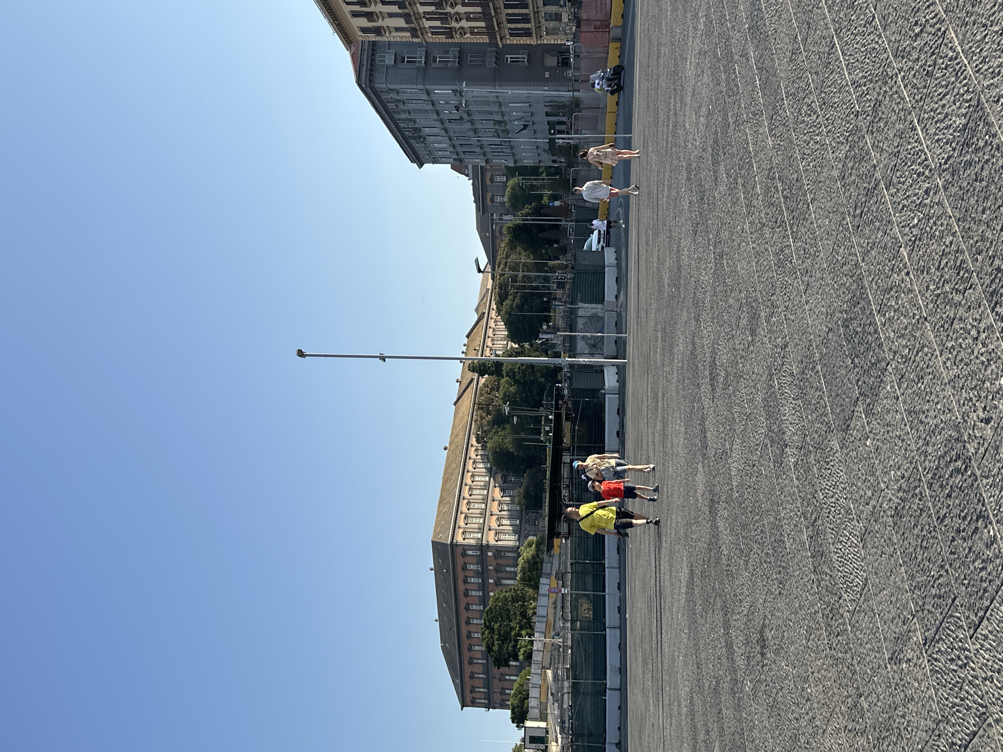 This image captures a sunny day in Piazza del Plebiscito, Naples, Italy. The foreground is dominated by the large, gray stone pavement of the piazza. In the middle ground, a family of three, consisting of a man in a yellow t-shirt, a woman in a light-colored outfit, and a child in a red t-shirt, walks across the piazza. Behind them, another couple, dressed in casual summer attire, walks away from the camera. To the right, a person on a scooter moves past.
In the background, the grand facade of the Palazzo Reale (Royal Palace) is visible, its reddish-brown brick and numerous windows stretching across the frame. Adjacent to the palace, construction fencing and barriers are in place, suggesting ongoing work or renovations. Further to the right, modern-looking apartment buildings line the edge of the piazza. The sky above is a clear, bright blue, indicating a cloudless day. A lamppost stands tall in the center of the frame.