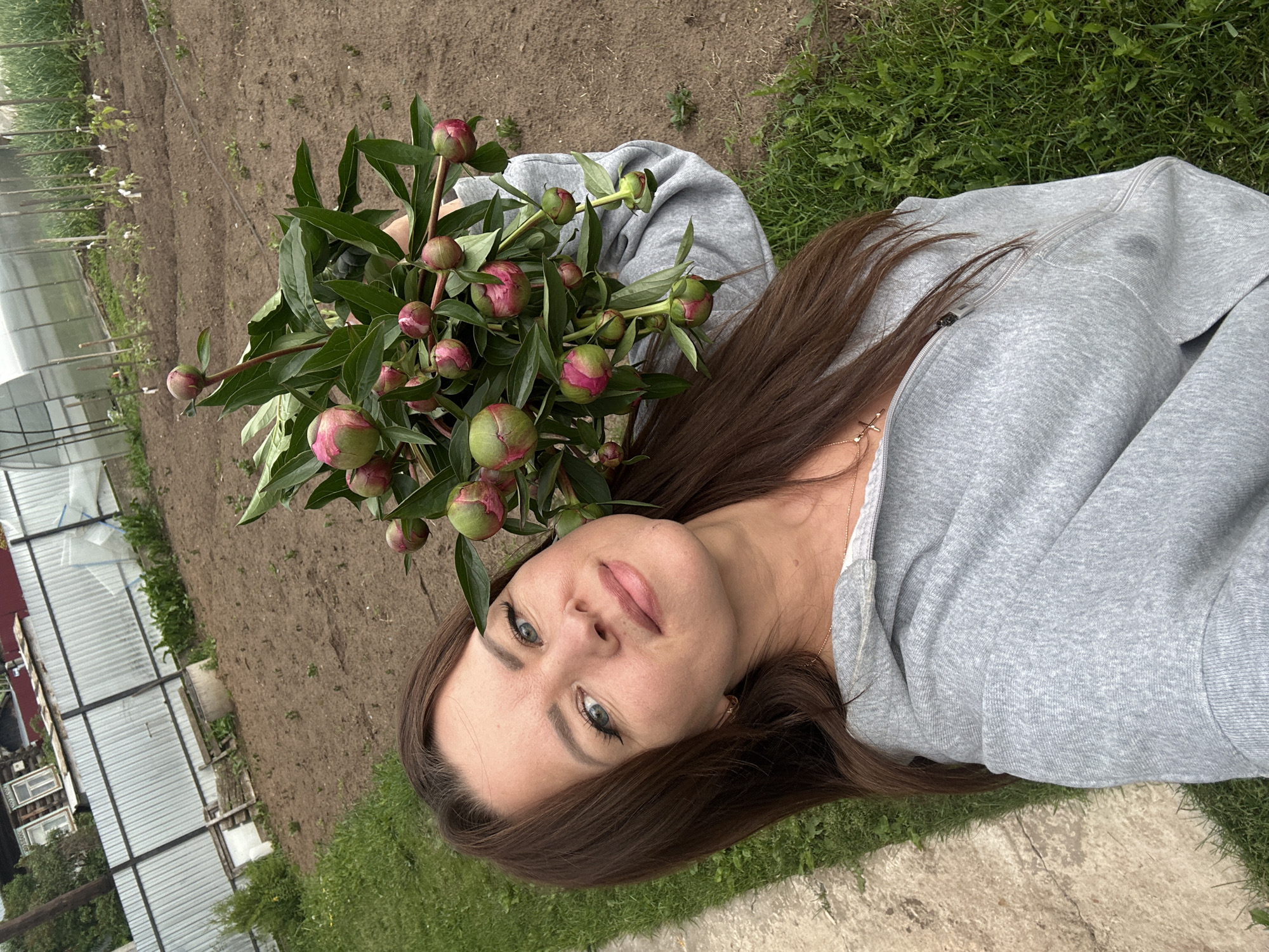 A forensic examination of the image reveals a female individual in a semi-rural setting, identified by the user as the city of Chaikovsky, Russia. The woman, appearing to be an adult, has long brown hair, blue eyes with dark eyeliner, and light-colored lipstick, and is looking directly at the camera with a subtle smile. She is wearing a grey hooded zip-up sweatshirt and a gold-colored necklace with a cross pendant. Her right arm and shoulder are visible, holding a substantial cluster of green peony buds with visible pink coloration on their tips, along with their associated green foliage.
The background consists of a cultivated garden or field. To the immediate left of the woman, a narrow concrete path runs vertically, bordered by green grass. The dominant background element on the right side is a large expanse of tilled brown earth, indicating recently prepared or planted garden beds, with some small green plants visible in sparse rows. In the mid-ground, a corrugated metal fence separates the garden area from what appears to be a residential structure – a wooden house with a red roof partially visible behind the fence on the left side. Also in the mid-ground, near the fence on the left, is a concrete well structure with some wooden planks beside it. Further in the background, to the right, a greenhouse with a transparent roof is present, alongside more rows of planted crops supported by vertical stakes, and a section of taller green crops. The overall environment suggests an agricultural or home gardening context within a residential or rural area.