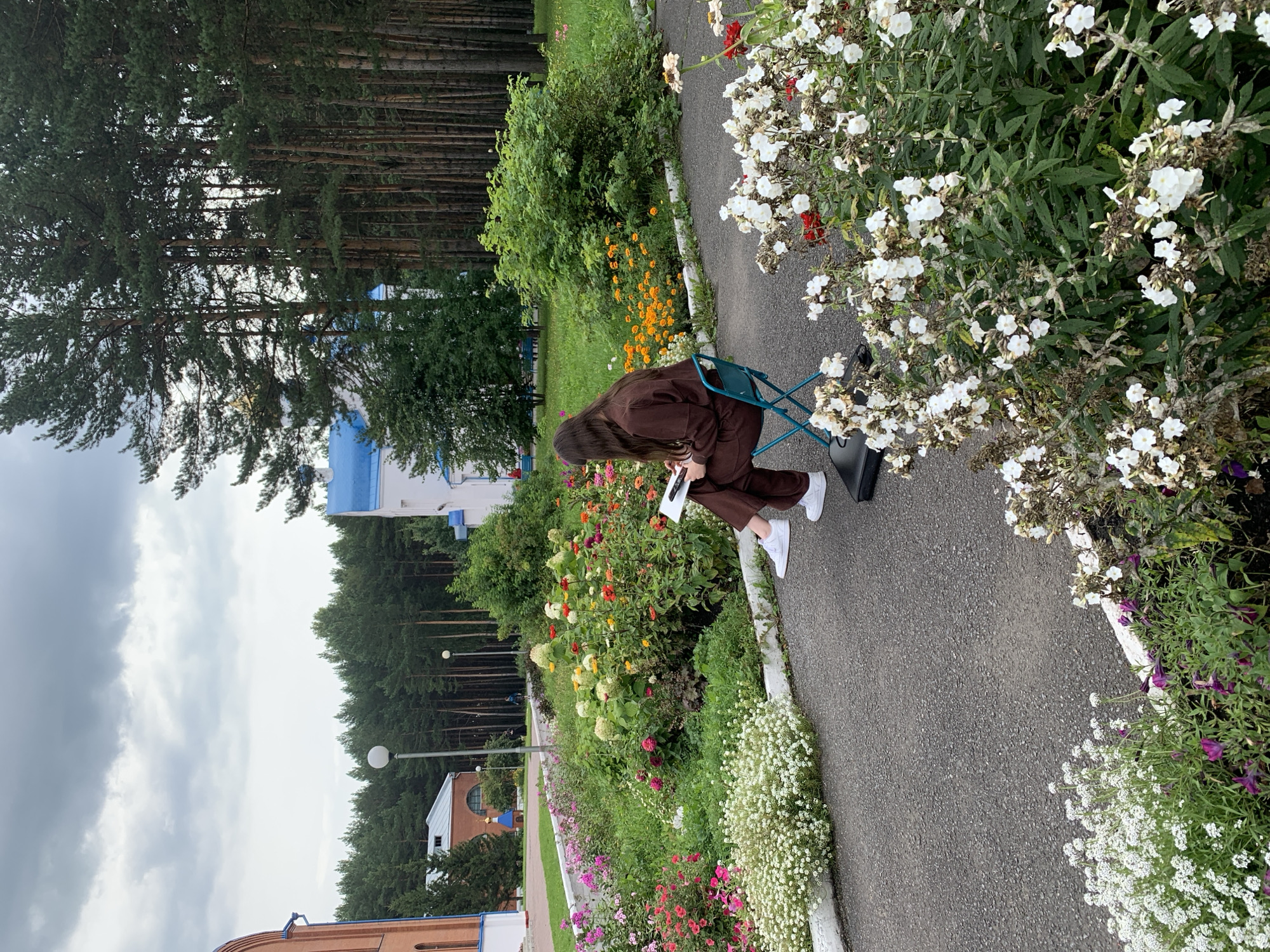 A person sits on a teal folding chair in front of a flowerbed, facing away from the camera. They are wearing a brown hooded jumpsuit and white sneakers, and are holding a tablet and stylus, appearing to be drawing or working on the device. The flowerbed contains a variety of colorful flowers, including pinks, reds, oranges, yellow, and white blossoms, interspersed with green foliage. A paved walkway runs alongside the flowerbed. In the background, tall pine trees form a dense forest. Several buildings with brick and white facades, and blue roofs, are visible through the trees. A street lamp stands near the walkway. The sky is overcast with grey clouds. The location is the territory of the St. George Monastery in the city of Prokopyevsk, Russia.