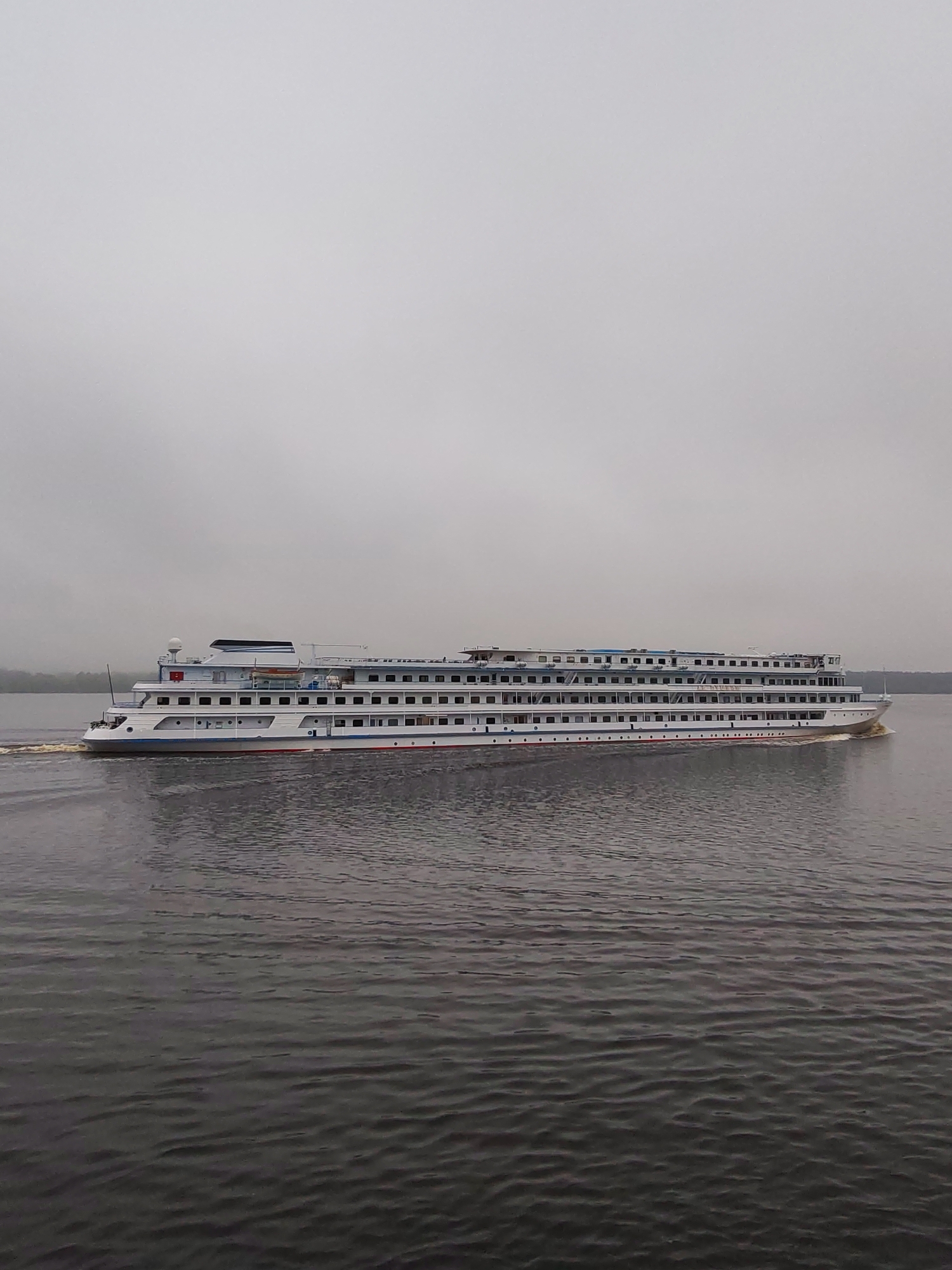 A long, multi-decked river cruise ship is sailing on a large body of water. The ship is predominantly white, with rows of windows on each deck and red and blue stripes along its hull. The water is dark and rippled. The sky is overcast and grey, suggesting a cloudy day. The location is Unknown, Unknown.