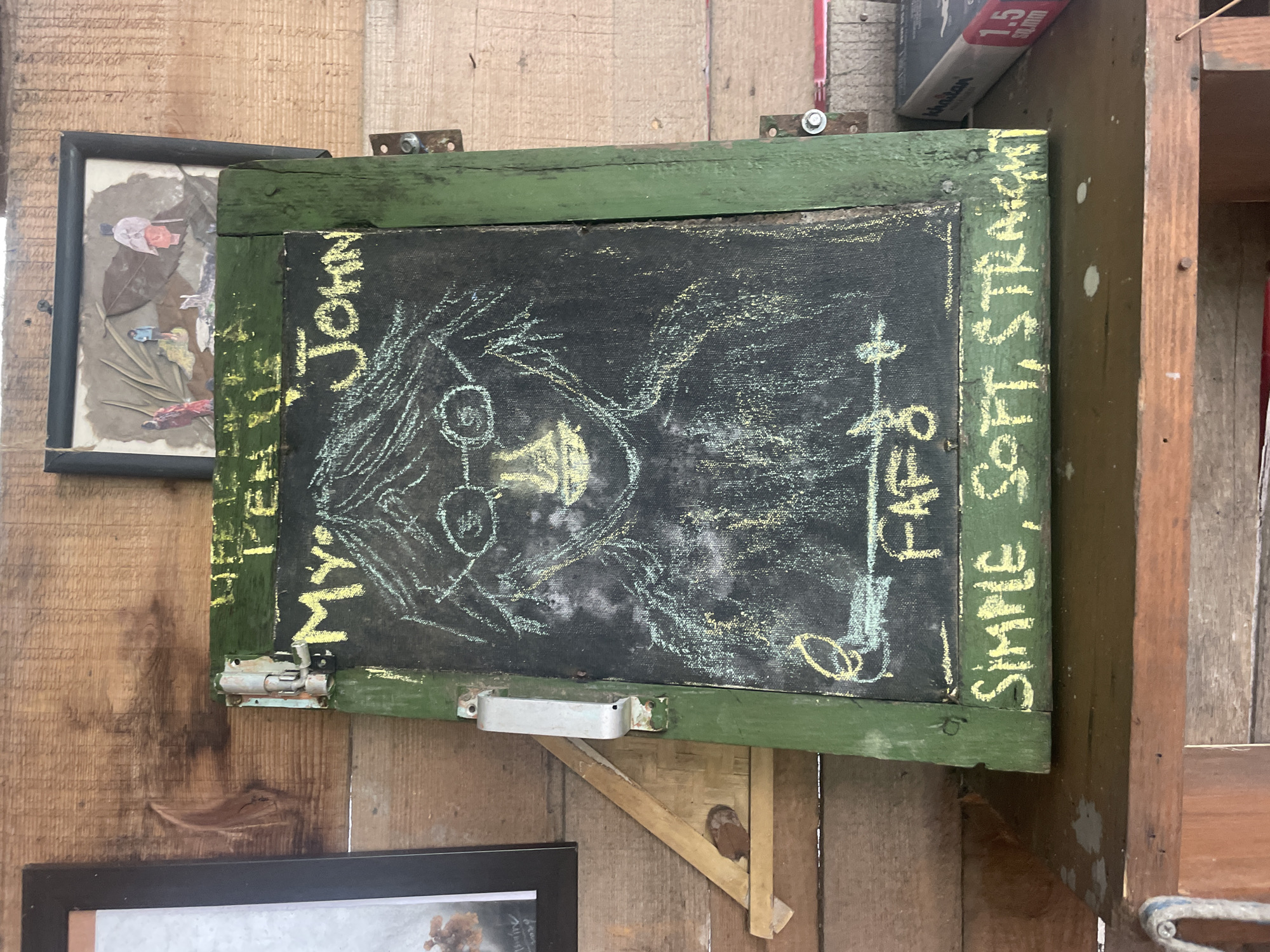In Bhagsu, India, a rustic wooden shelf displays a vintage chalkboard painted green on its frame, featuring a chalk drawing of what appears to be John Lennon. The drawing, rendered in yellow chalk, depicts Lennon with circular glasses and tousled hair. Above the portrait, the words "HAPPY PEACE LOVE" are scrawled in yellow chalk, followed by "MY JOHN". Below the portrait, the word "FARGO" is written, and further down, "SIMPLE, SOFT, STRAIGHT" is inscribed in yellow chalk. The chalkboard is mounted on a wooden wall, with a small framed artwork of three figures above it. A metal latch secures the chalkboard to the frame. To the right, a box of "KHAITAN" branded items, possibly electrical equipment, is visible on the shelf.