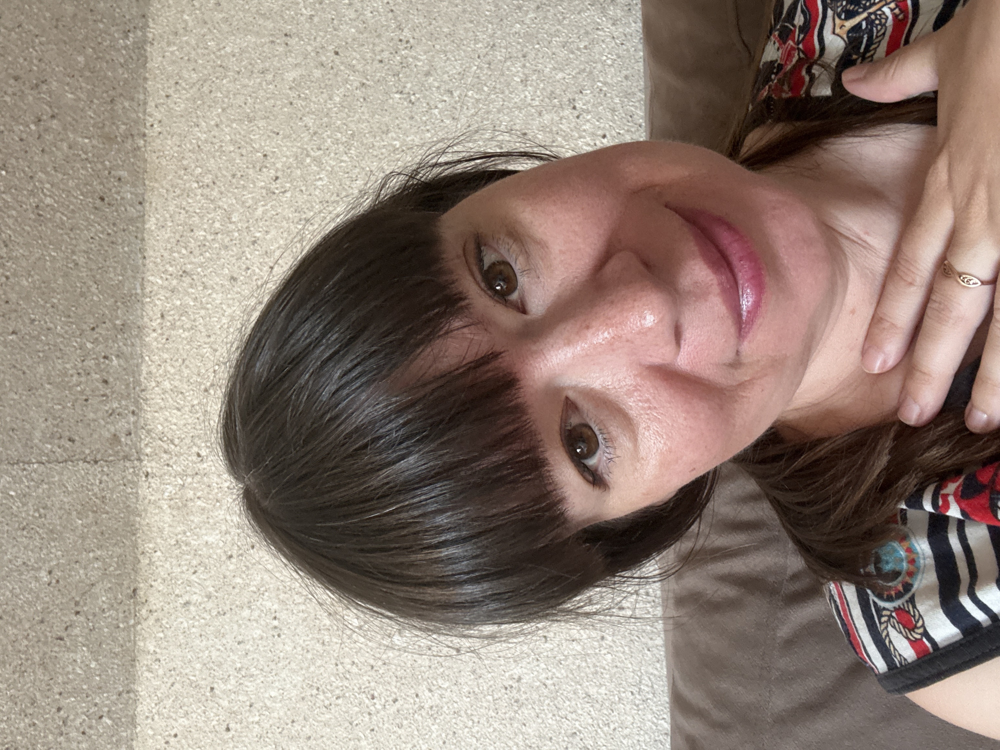 A close-up, selfie-style photograph shows a woman with dark brown hair and bangs. She is looking at the camera with brown eyes and a slight smile. Her right hand is resting on her neck, and she is wearing a patterned shirt with black, white, red, and blue stripes. The background appears to be a textured wall, likely inside a building. No specific location within Vetluzhsky, Russia can be determined from the image.