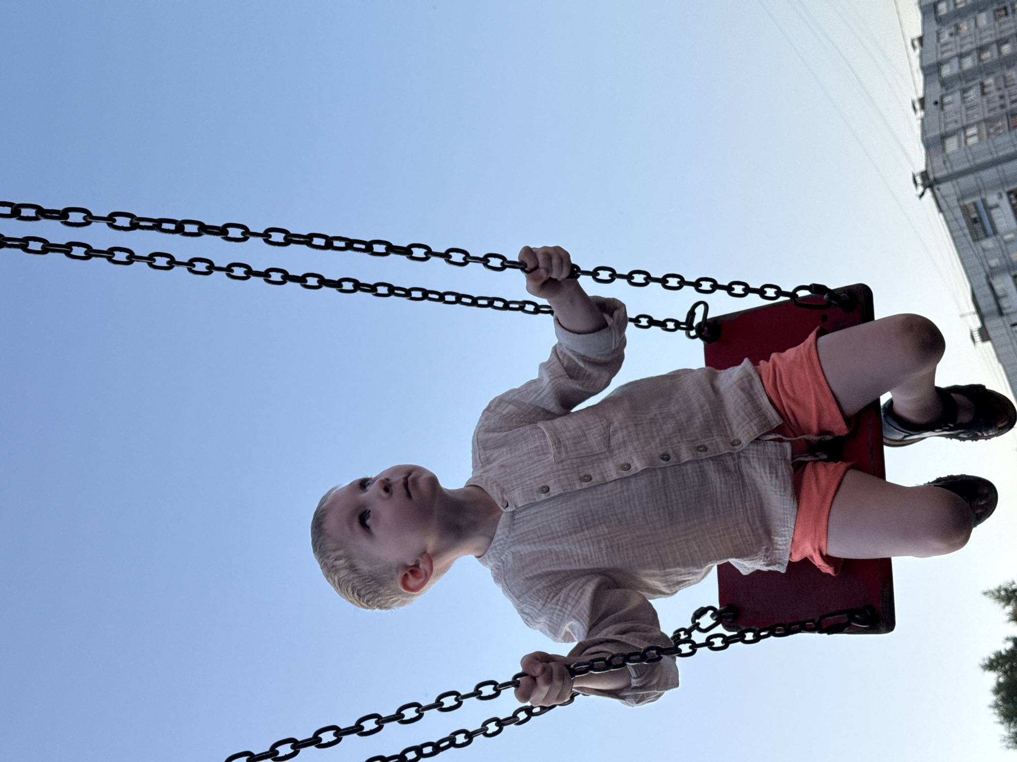 A child is on a swing, viewed from below. The child wears a light-colored, buttoned shirt and orange shorts, with black sandals on their feet. The swing has metal chains and a red seat. The sky is blue. A tall building is visible in the background. The location is possibly Chaikovsky, Russia.