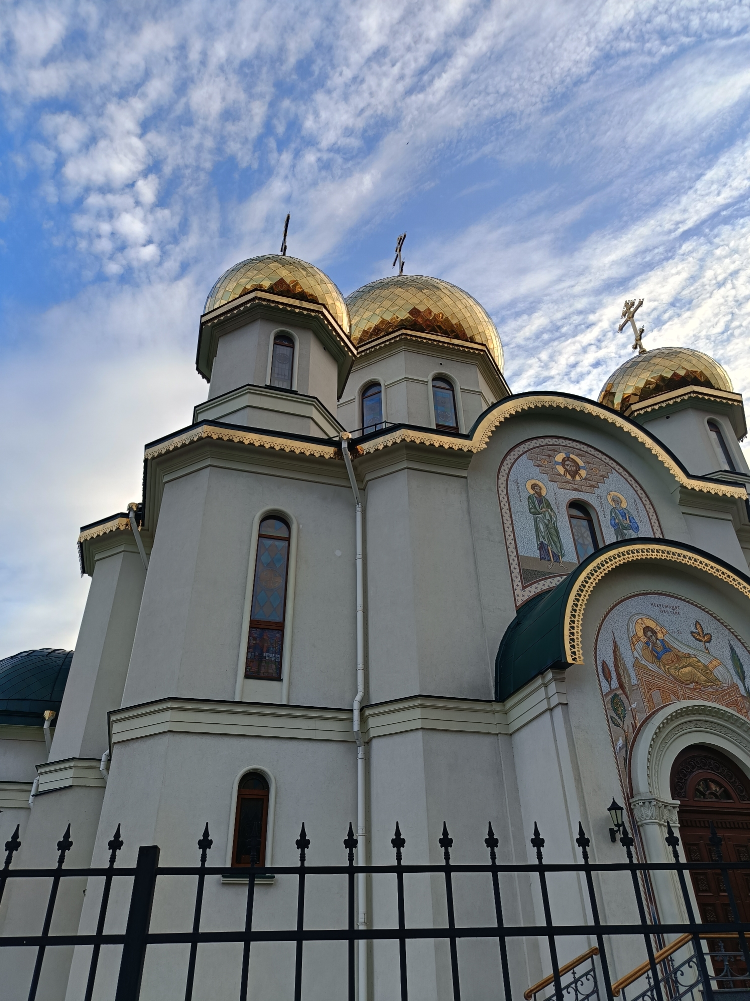 The image shows an Eastern Orthodox church with golden domes against a cloudy blue sky. The church has a light-colored facade adorned with mosaics depicting religious figures. A black metal fence is in the foreground. This is the Church of the Resurrection of Christ in St. Petersburg, Russia.
