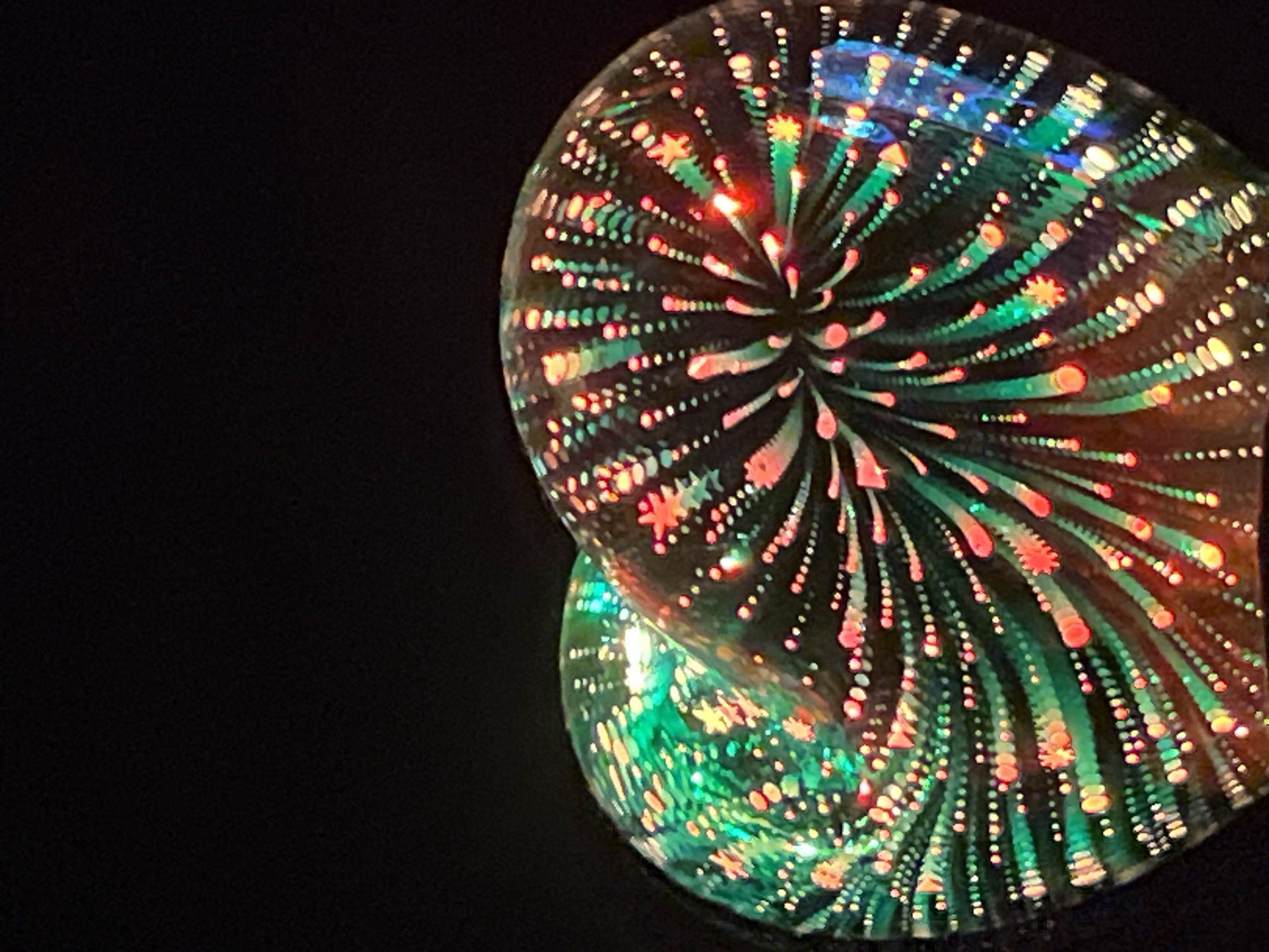 In an unknown location, two translucent glass orbs are illuminated from within, creating a mesmerizing display of light and color against a black background. The orbs are positioned close together, with the left orb exhibiting predominantly green and yellow hues, while the right orb displays a mix of red, pink, and blue light. Both orbs feature intricate patterns of small, bright dots and star-like shapes that swirl outwards from a central point, resembling abstract fireworks or nebulae. The lighting within the orbs casts a soft glow on their curved surfaces, and reflections of the lights are visible on the outer edges of the glass. The overall impression is one of vibrant, dynamic illumination contained within delicate glass spheres.