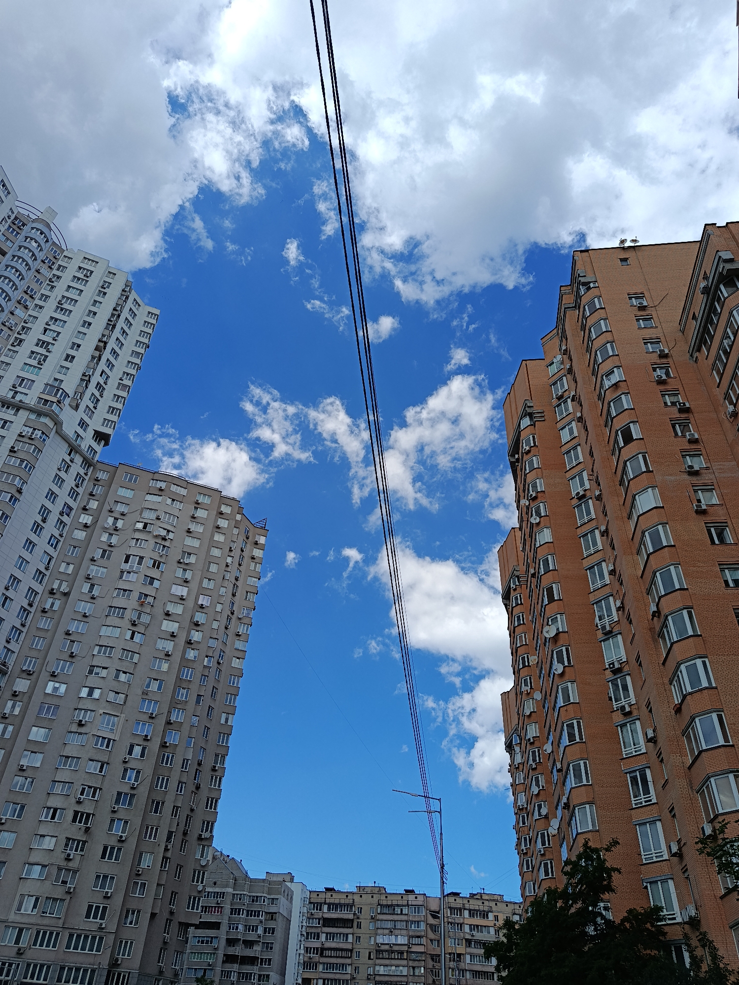 This is a low-angle, full shot of a city skyline in Kyiv, Ukraine, dominated by a bright blue sky with scattered white clouds. On the left side of the frame, a tall, modern residential building with a white facade and curved balconies stands prominently. To its right, a slightly shorter building with a grey facade and numerous windows and air conditioning units is visible. On the right side of the image, a very tall residential building constructed of orange brick is featured, its facade also covered with windows and AC units. Several power lines stretch across the upper portion of the frame, dividing the sky. In the background, more residential buildings of varying heights and architectural styles can be seen. The overall scene depicts a dense urban environment with a clear sky.