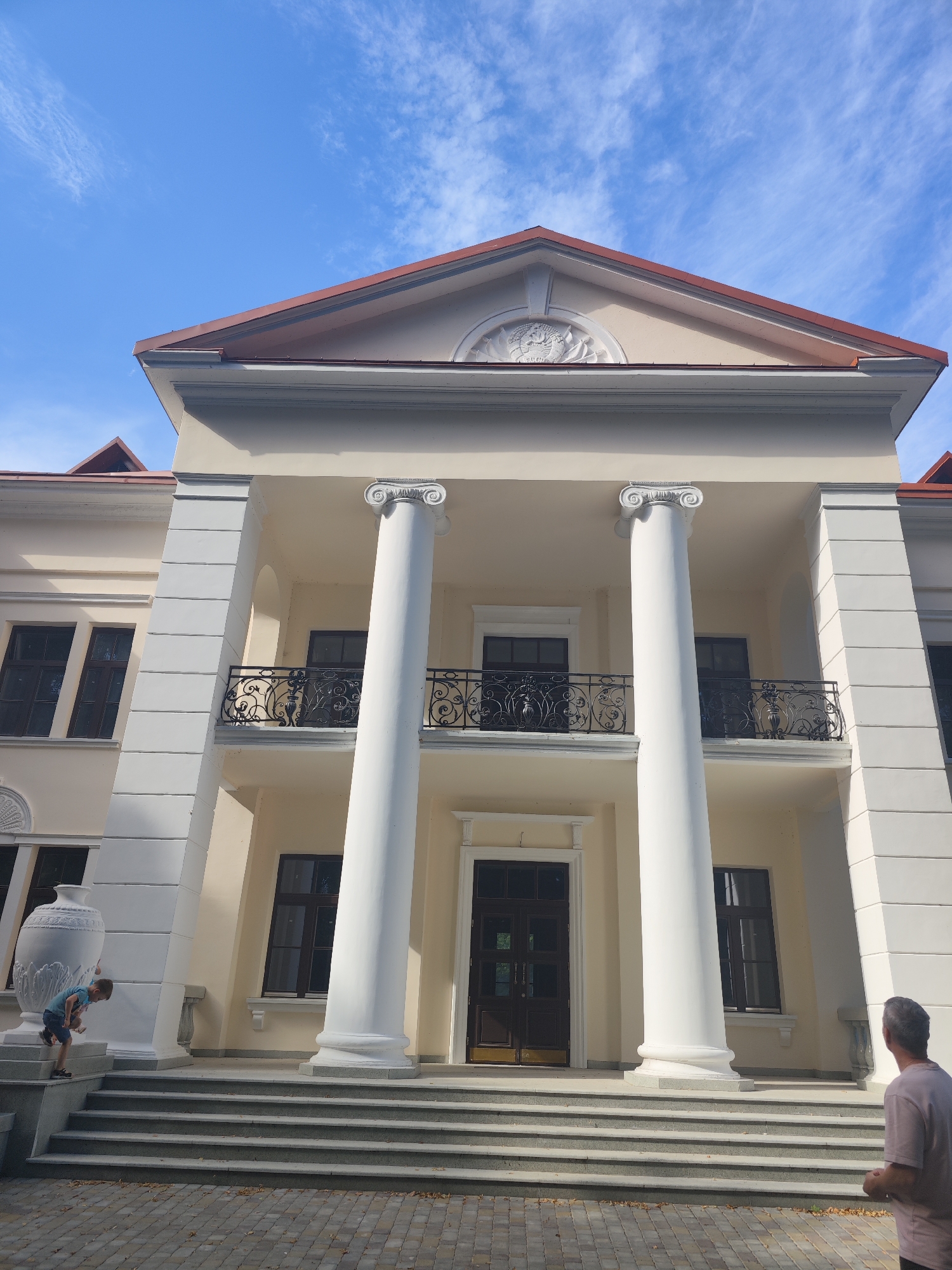 The image depicts the facade of a pale yellow, neoclassical-style building with a red-tiled roof and white columns. A young boy in a blue shirt and shorts is on the steps of the building, leaning against a large white urn. A man in a light-colored t-shirt is standing on the right side of the image, looking towards the building. The sky above is blue with scattered white clouds. The building appears to be a historical or public structure, possibly a museum or government building, and the overall scene suggests a warm, sunny day. This building is located in the city of Slonim, Belarus.
