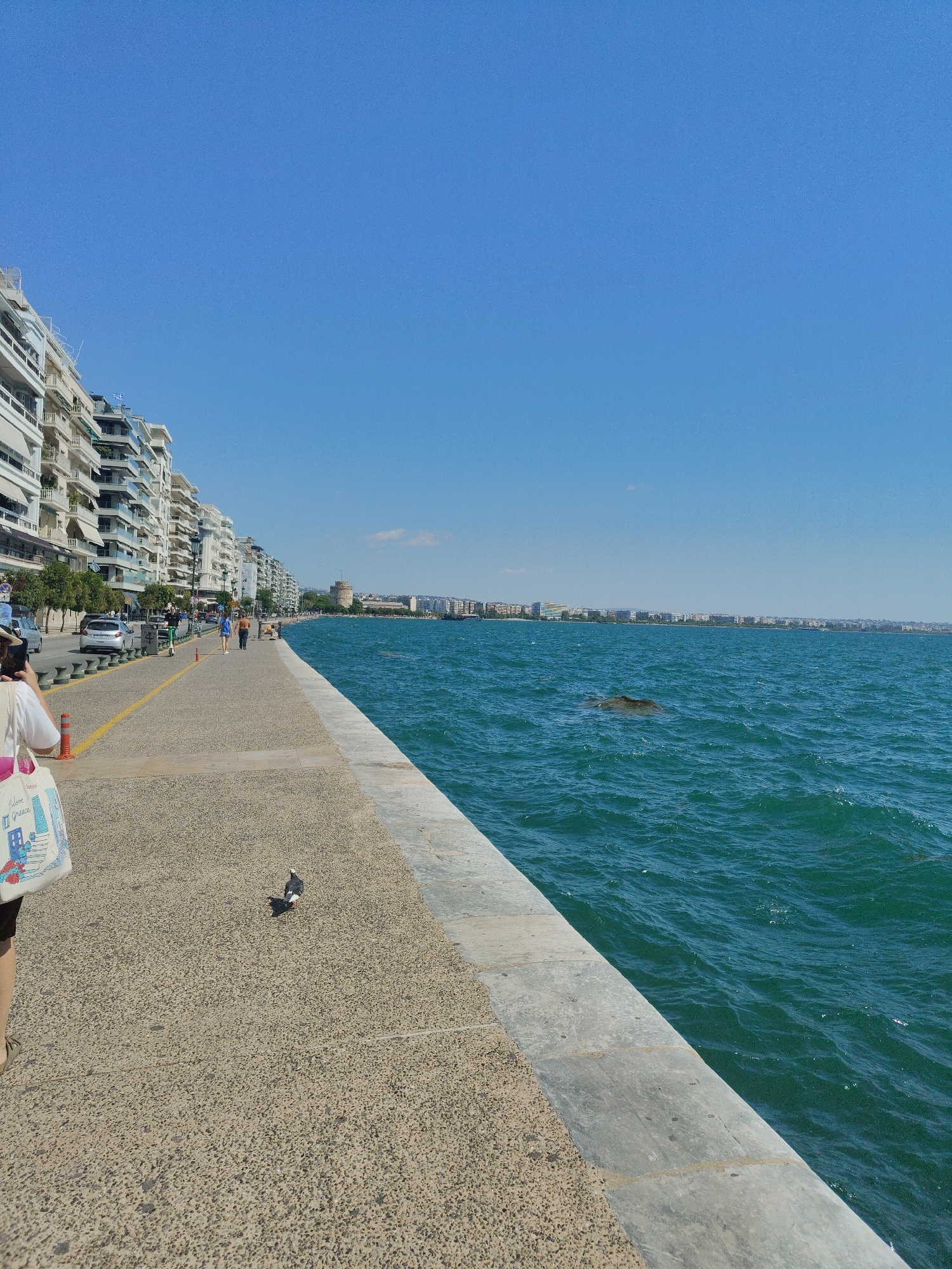 A wide-angle, eye-level, full shot captures a sunny day scene along the waterfront promenade in Thessaloniki, Greece. The clear blue sky dominates the upper portion of the frame, meeting the turquoise waters of the sea. A paved walkway, bordered by a low, light-colored stone wall, runs along the coast. On the left side of the walkway, a row of multi-story apartment buildings with white facades and balconies lines the street. Cars are parked along the street, and pedestrians are seen walking along the promenade.
In the mid-ground, two people, a woman in a blue top and a man in a brown shirt, walk away from the camera. Further down the promenade, the iconic White Tower of Thessaloniki is visible in the distance, a prominent landmark. In the sea, a dark, submerged object, possibly a rock or debris, breaks the surface.
On the walkway in the foreground, a pigeon is captured mid-stride, looking to its left. To the left of the pigeon, a person's legs are visible, wearing dark shorts and brown sandals. They are holding a white tote bag with a colorful design, including the words "I love Greece" and an illustration of buildings. The person is positioned in the lower left corner of the frame, partially obscuring the view of the street. The overall atmosphere is one of a pleasant, active urban waterfront on a clear day.